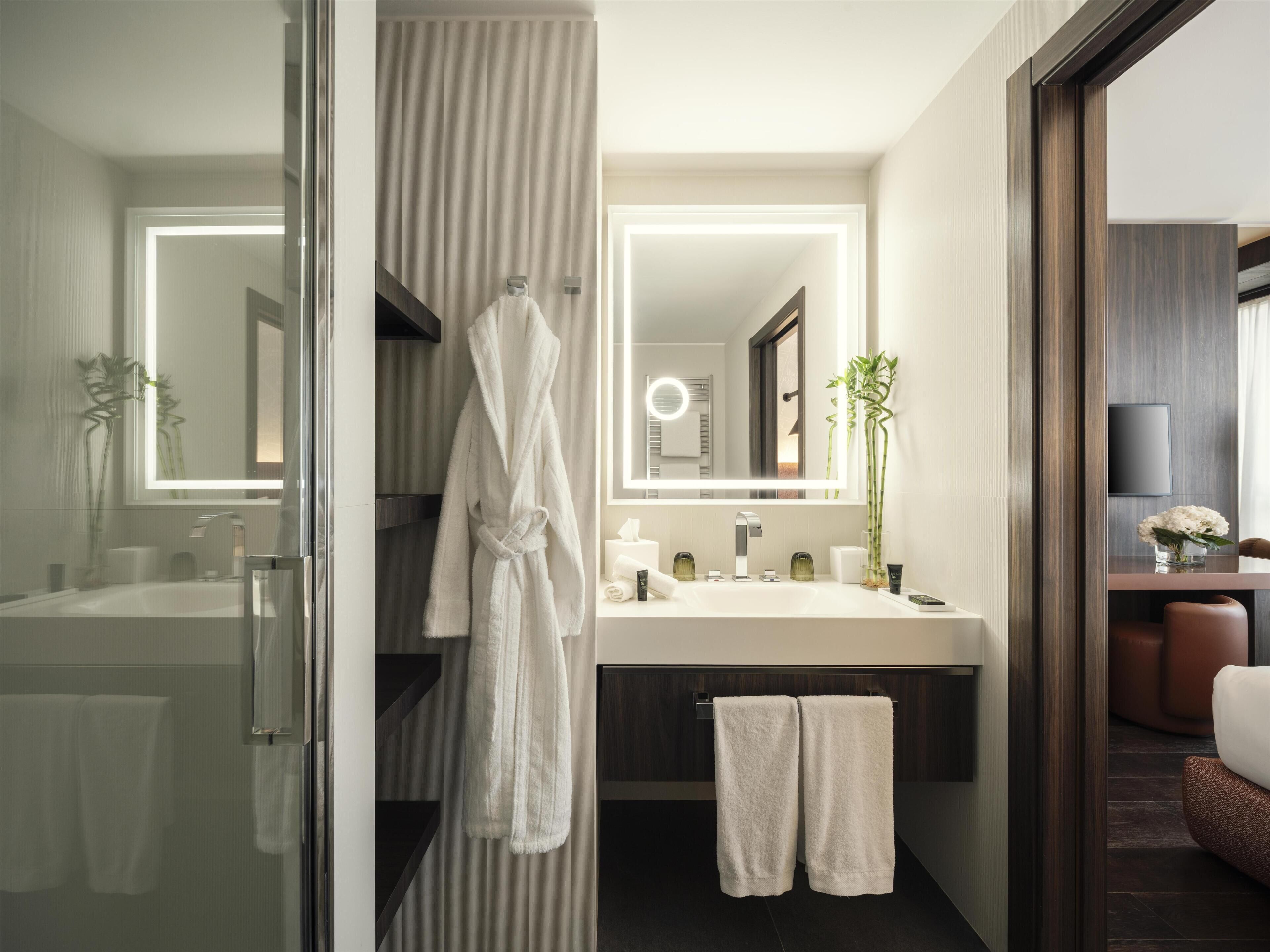 A modern bathroom with a glass shower enclosure, a white bathtub, a large mirror with integrated lighting, a vanity with a dark countertop, and a seating area with a brown leather chair.