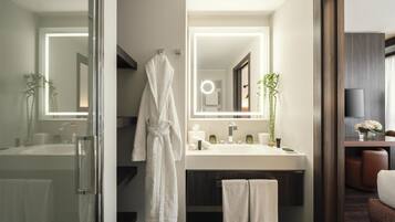A modern bathroom with a glass shower enclosure, a white bathtub, a large mirror with integrated lighting, a vanity with a dark countertop, and a seating area with a brown leather chair.