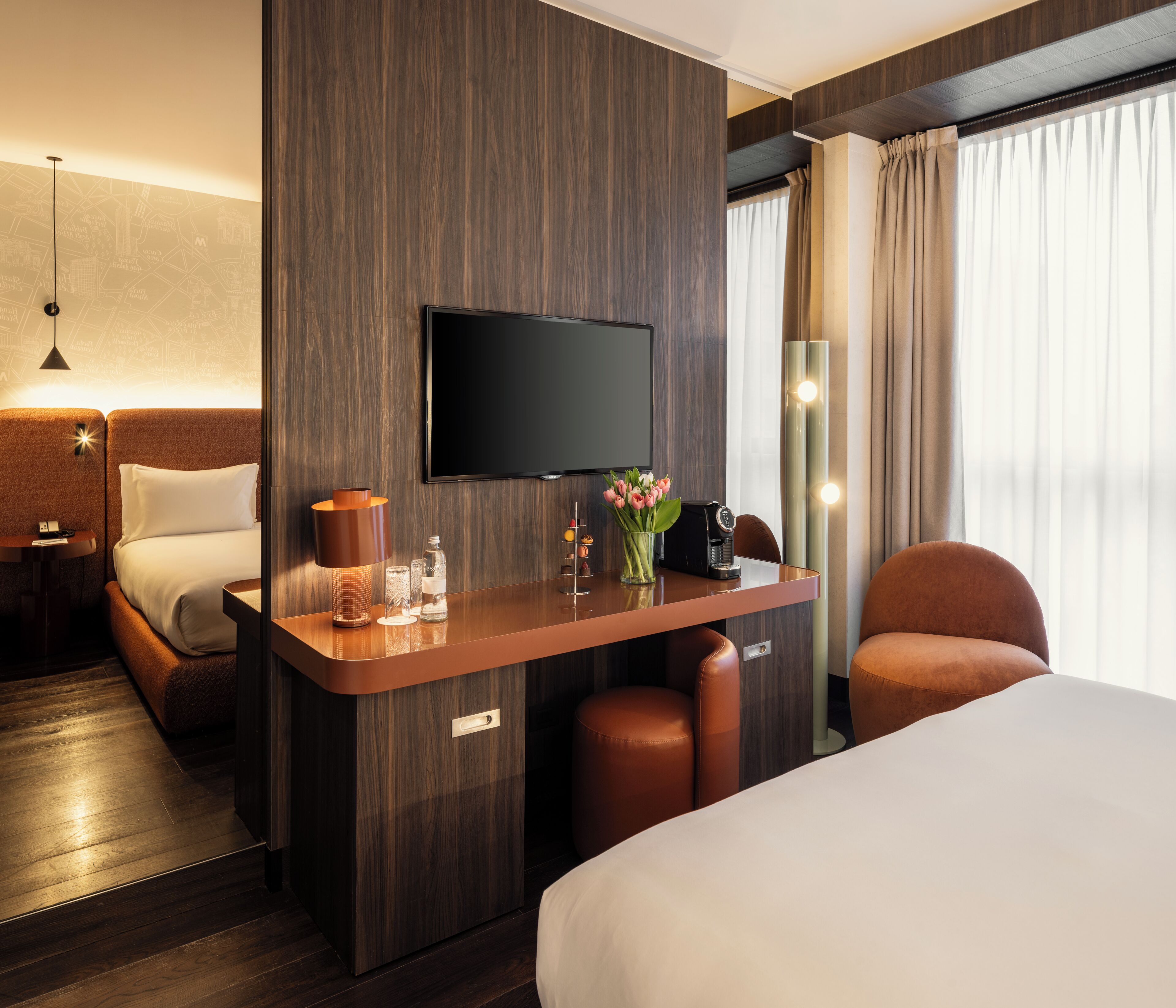 A hotel room with a bed, a desk with a flat-screen TV, a lamp, a vase with flowers, and a chair.