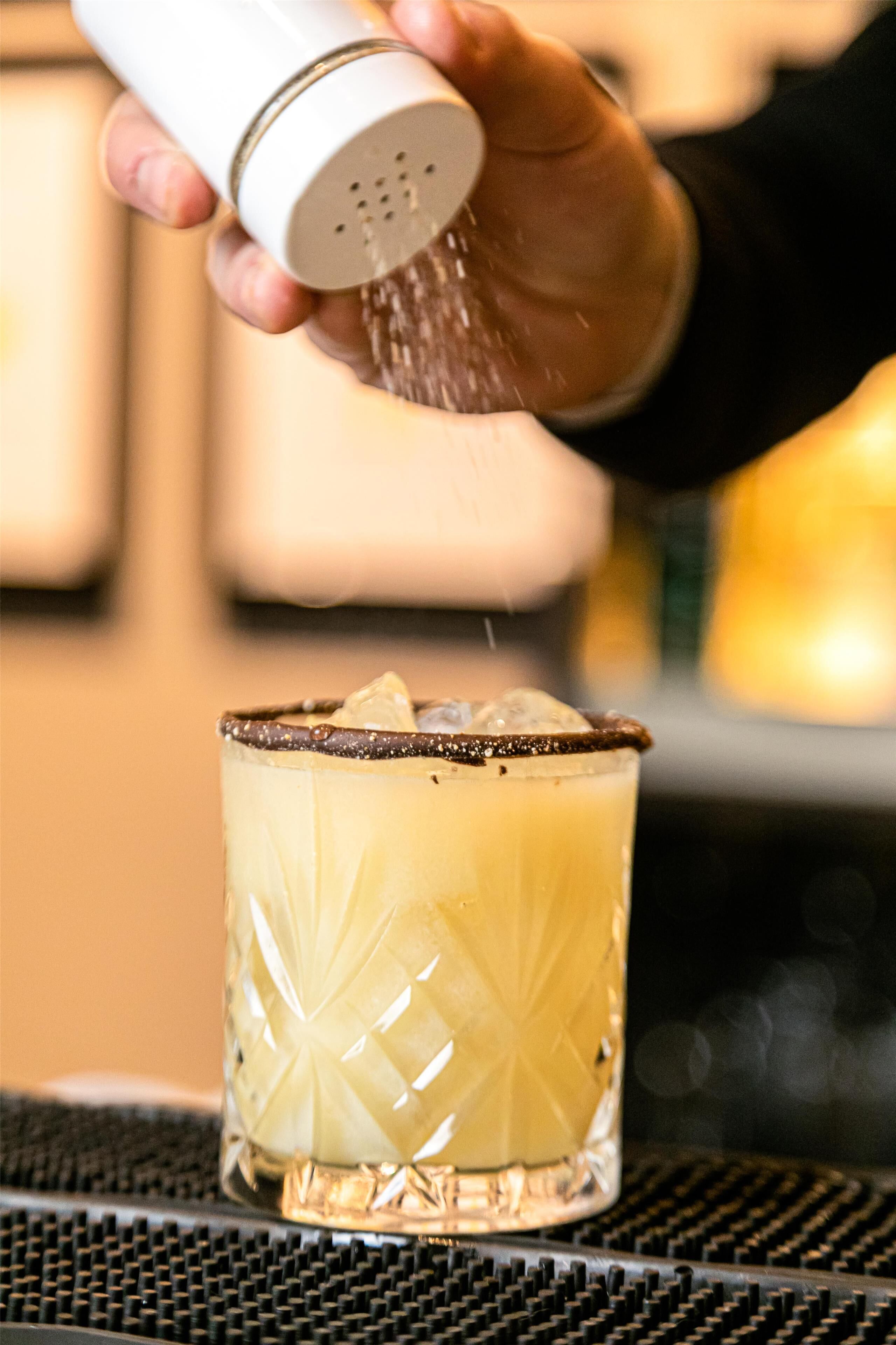 Een hand strooit een substantie over een glas met een patroon, geplaatst op een oppervlak met een raster.