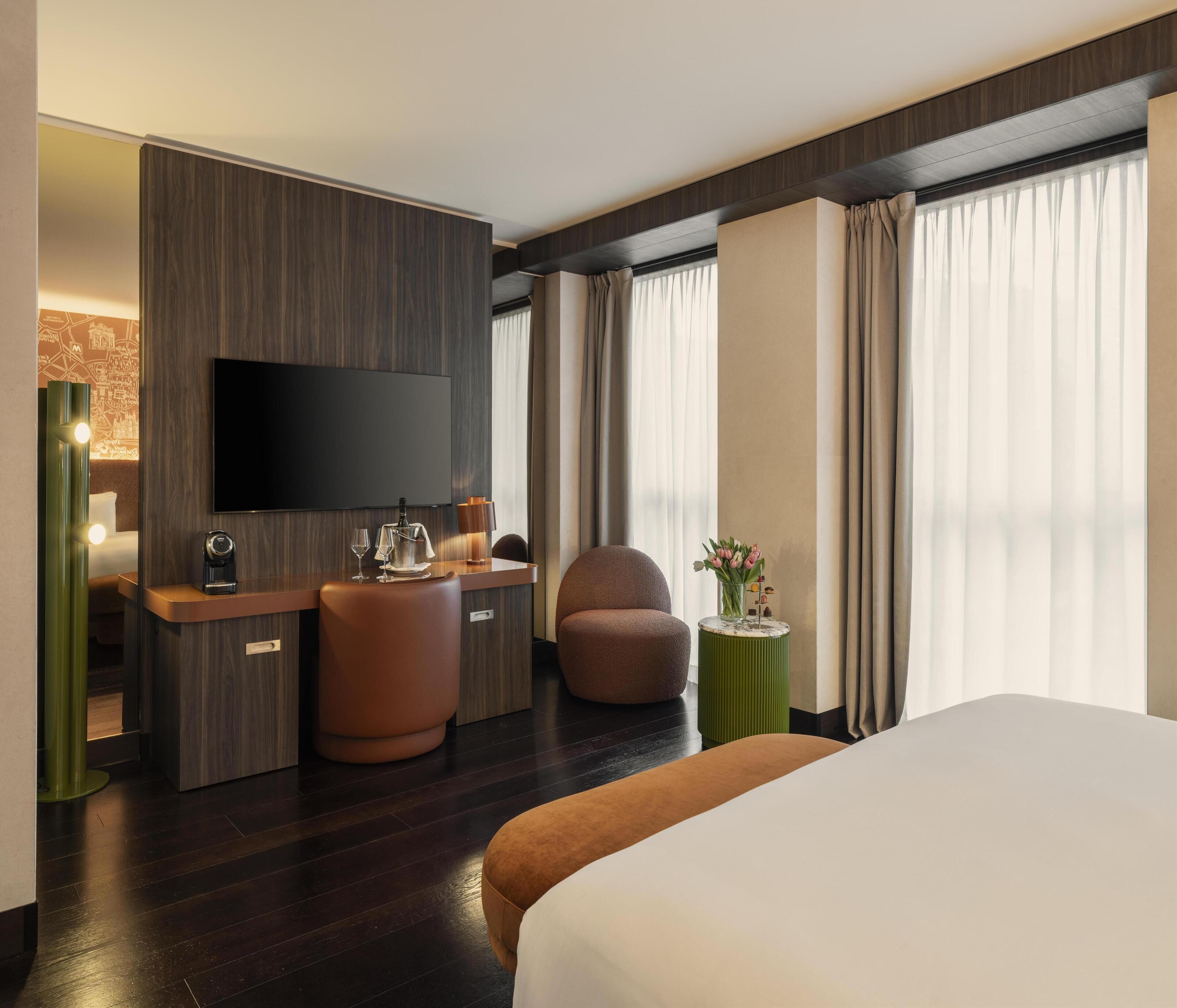 Hotelkamer met een bed, een bureau met flatscreen televisie, een stoel, een klein bijzettafeltje met een vaas, en een groot raam met gordijnen.