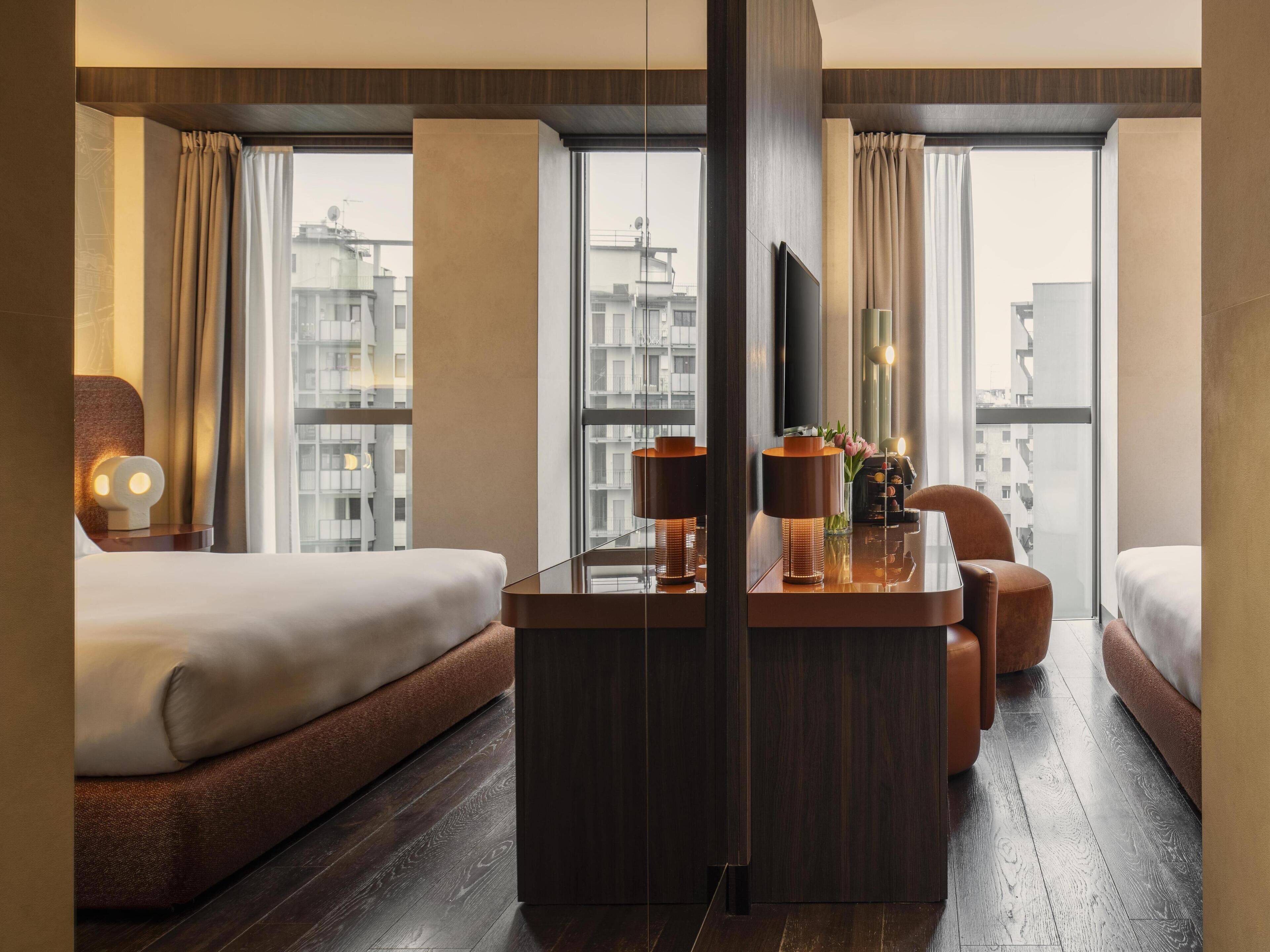 Quarto de hotel com duas camas, uma mesa de trabalho com luminária, televisão e vista para a cidade.