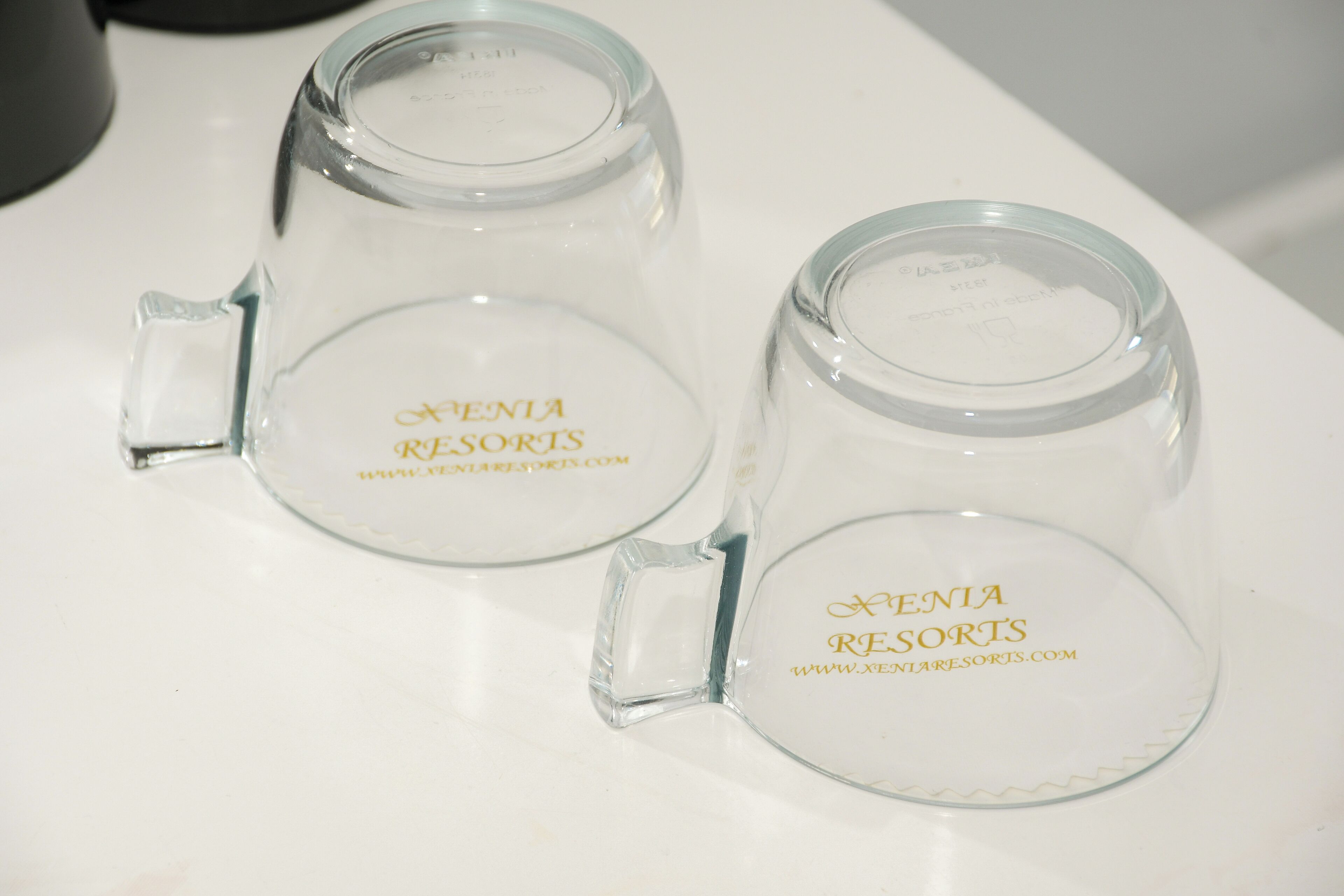 Dues tasses de vidre transparent amb mànecs, cadascuna amb l’etiqueta 'XENIA RESORTS' i una adreça web.