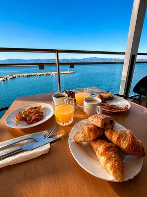 Desayuno con cruasanes, tocino, zumo de naranja y café sobre una mesa con vistas al puerto.