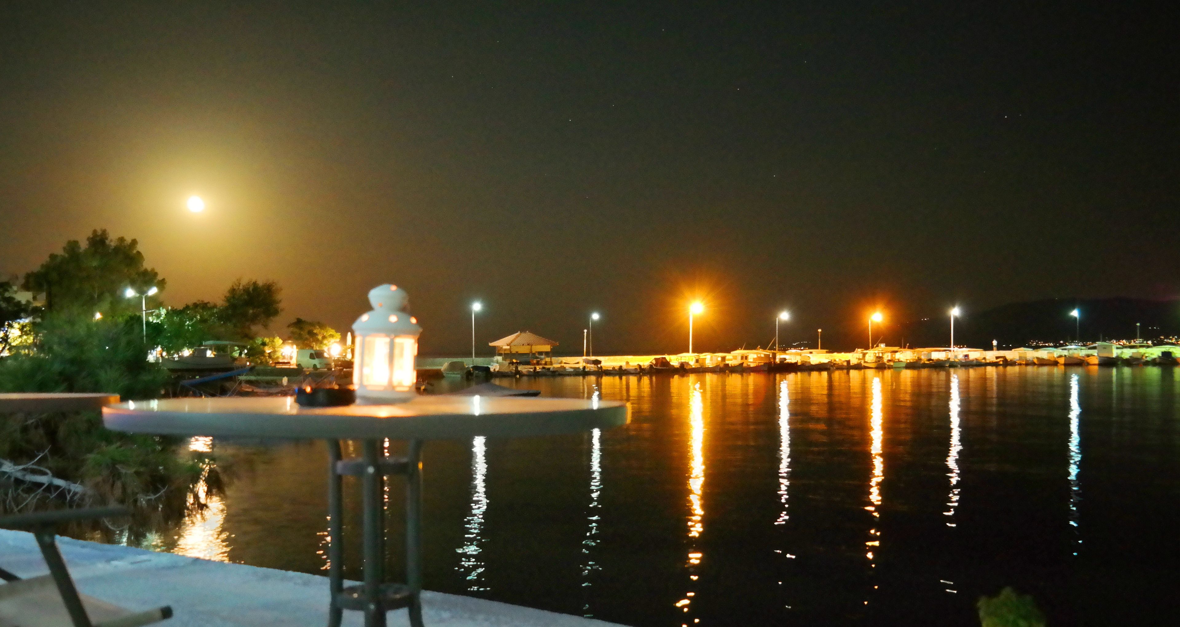 A nighttime view of a waterfront area with a table and chairs, a lantern, and a calm body of water reflecting lights.
