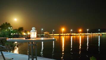 A nighttime view of a waterfront area with a table and chairs, a lantern, and a calm body of water reflecting lights.