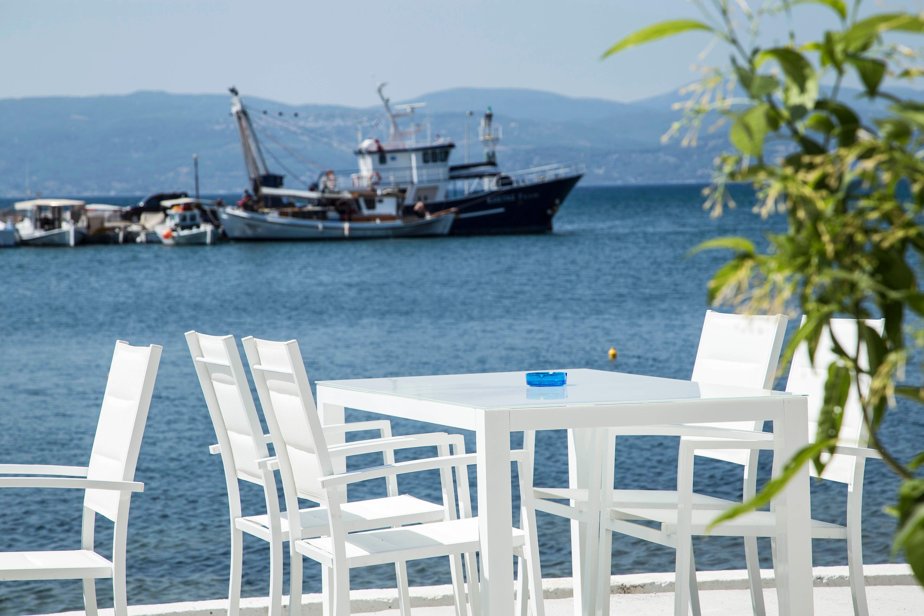 Una mesa con sillas blancas junto a un cuerpo de agua, un barco grande al fondo y una cadena montañosa en la distancia.