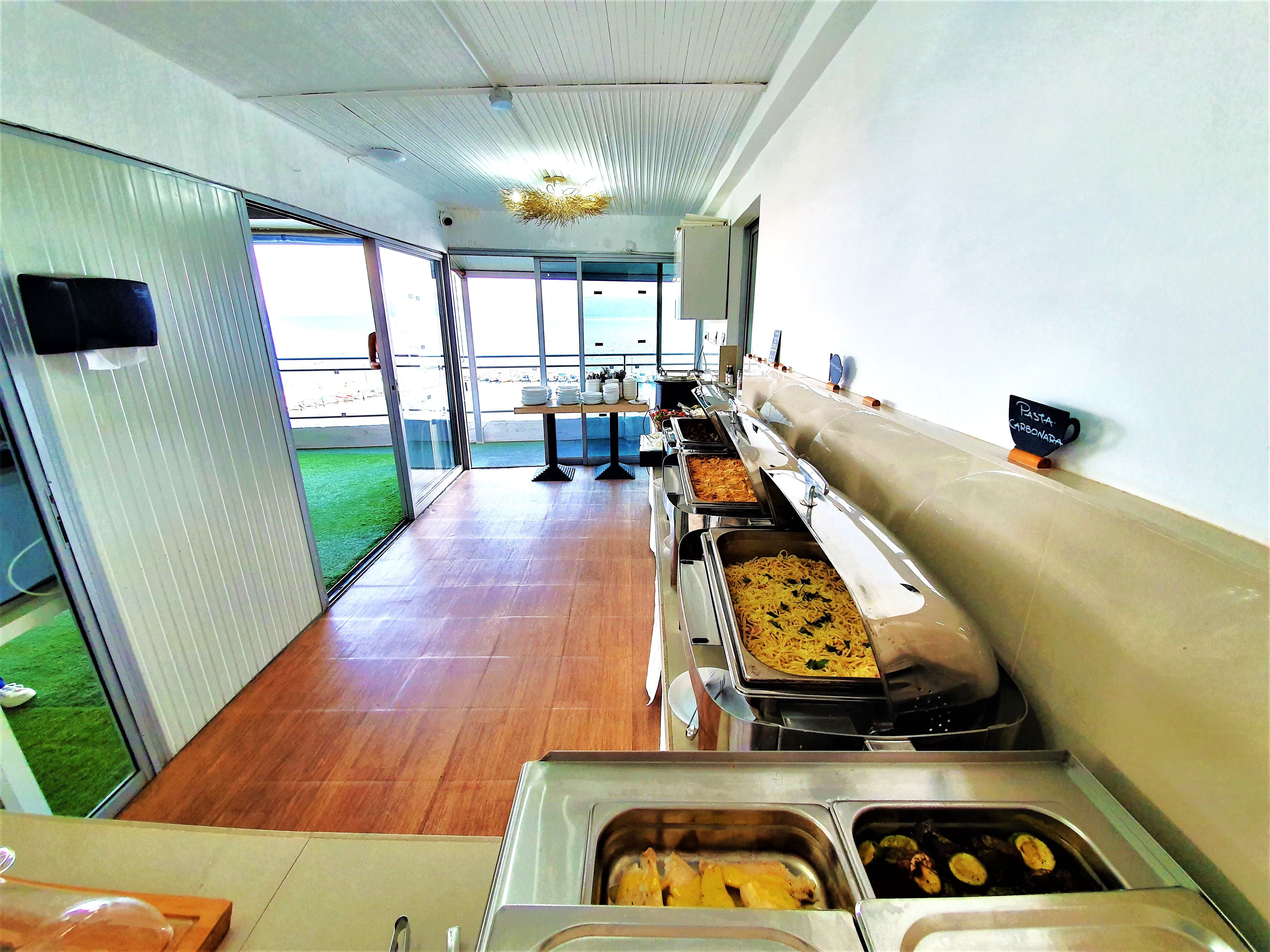 Un montaje tipo buffet con varias estaciones de comida, vistas al exterior a través de amplios ventanales y un suelo de baldosas.
