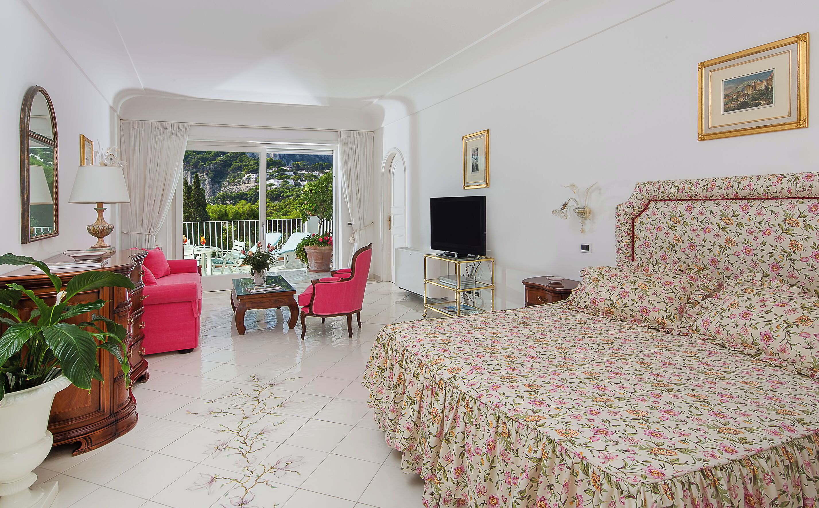 Habitación de hotel con cama, televisión, zona de comedor con sillas y vistas a zonas verdes.