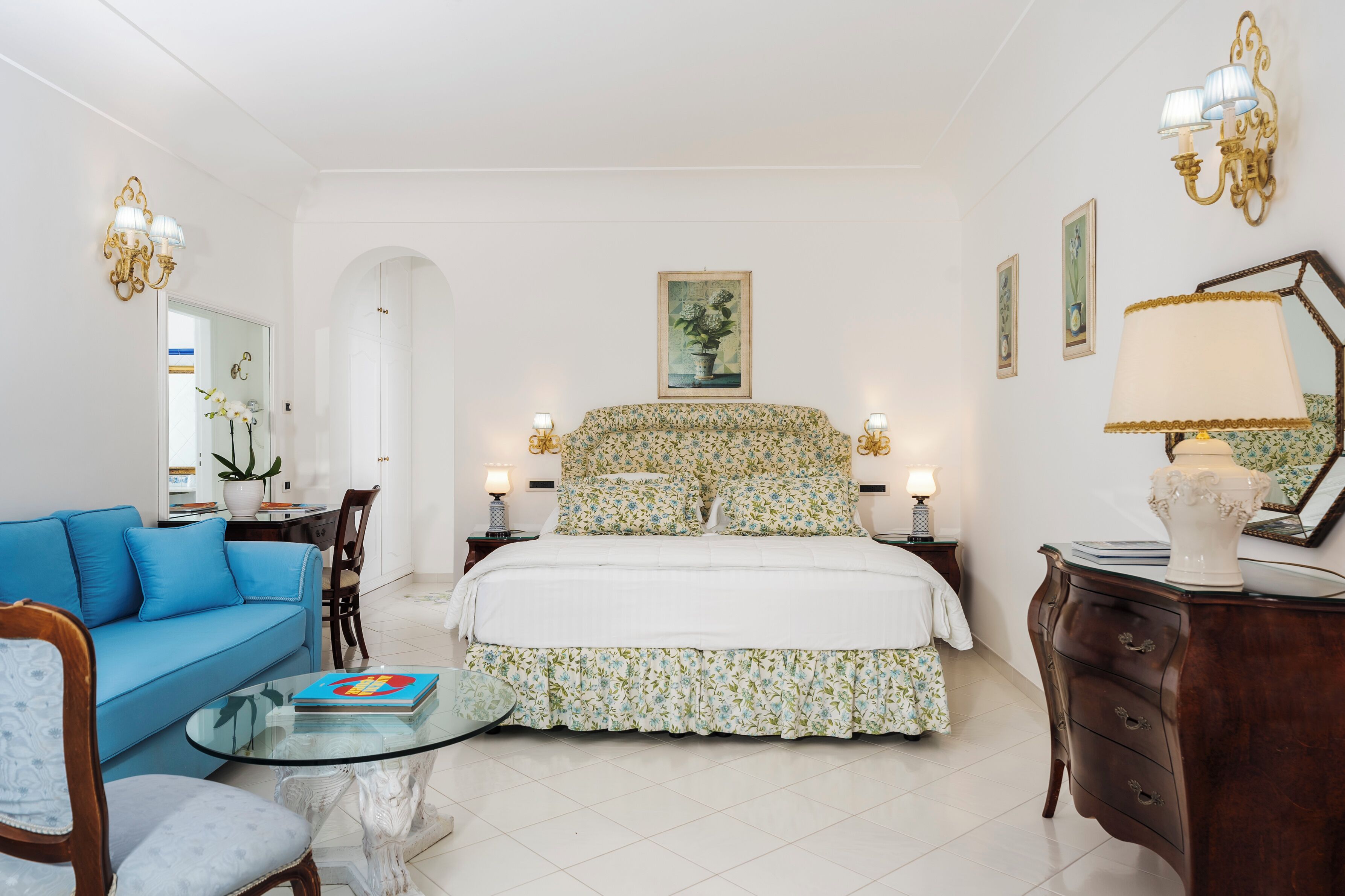 A bedroom with a bed, a blue sofa, a glass coffee table, and a dresser.