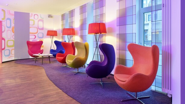 Ein modernes Interieur mit farbenfrohen, ungewöhnlich geformten Stühlen, die in einer Reihe angeordnet sind, einem violetten Teppich und einer Wand mit Muster.