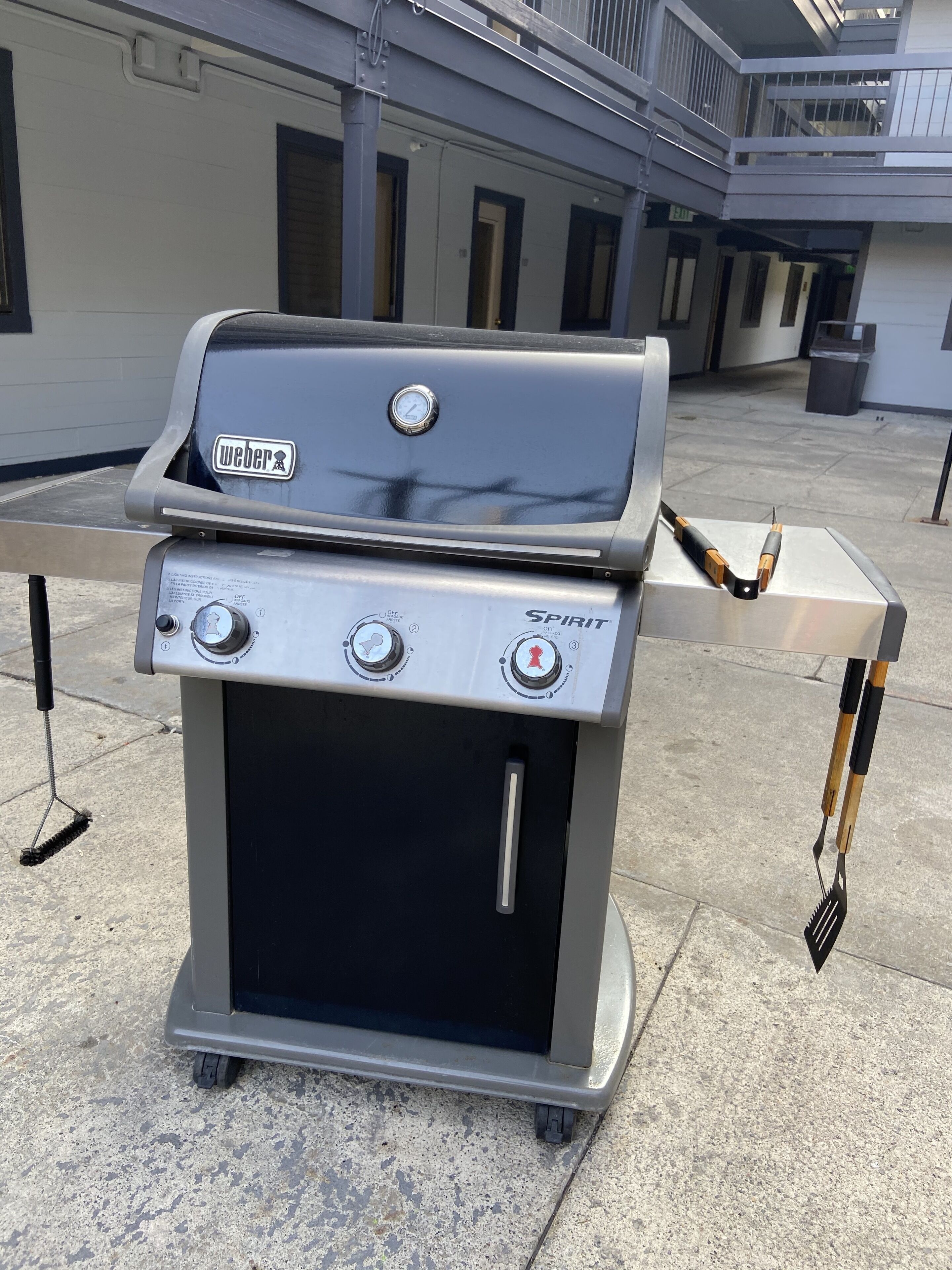 Un barbecue à gaz portatif Weber Spirit, doté d’un thermomètre numérique, de boutons de contrôle et d’une table d’appoint avec des ustensiles.