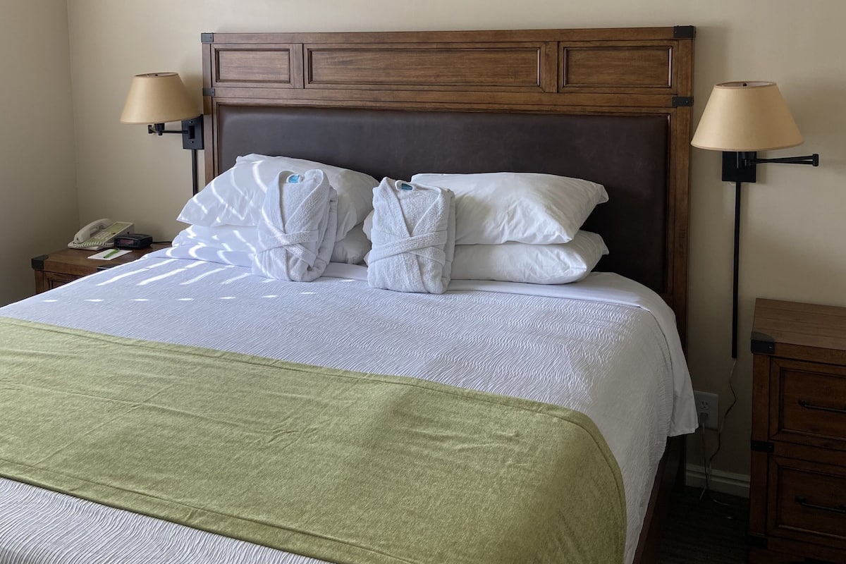 A neatly made bed with white linens and a green throw blanket, two bedside lamps, a wooden headboard, and a nightstand with a phone and a small table.