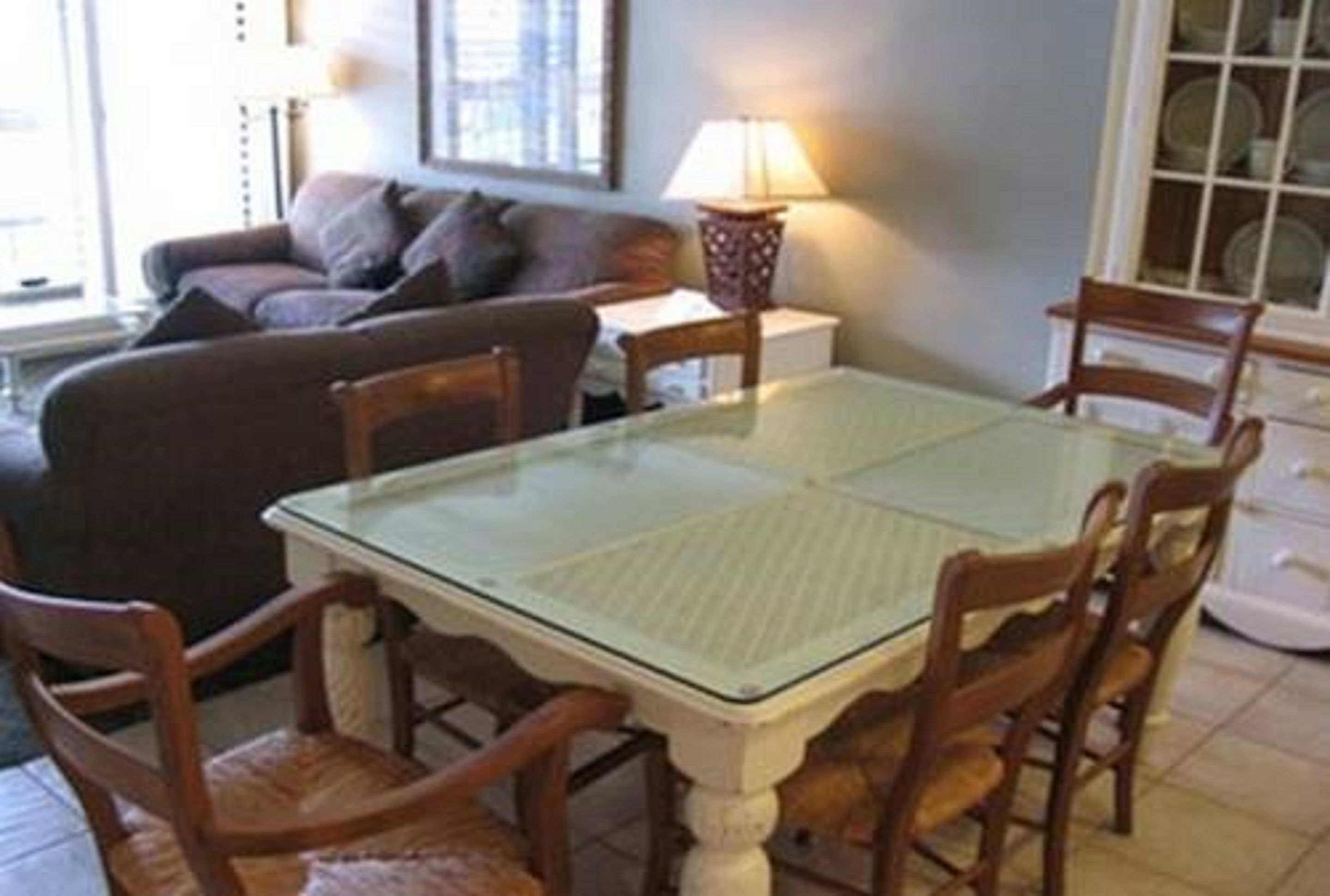 A dining area with a glass-top table and wooden chairs, a sofa, a lamp, and a window with blinds.