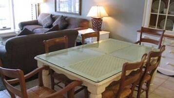 A dining area with a glass-top table and wooden chairs, a sofa, a lamp, and a window with blinds.