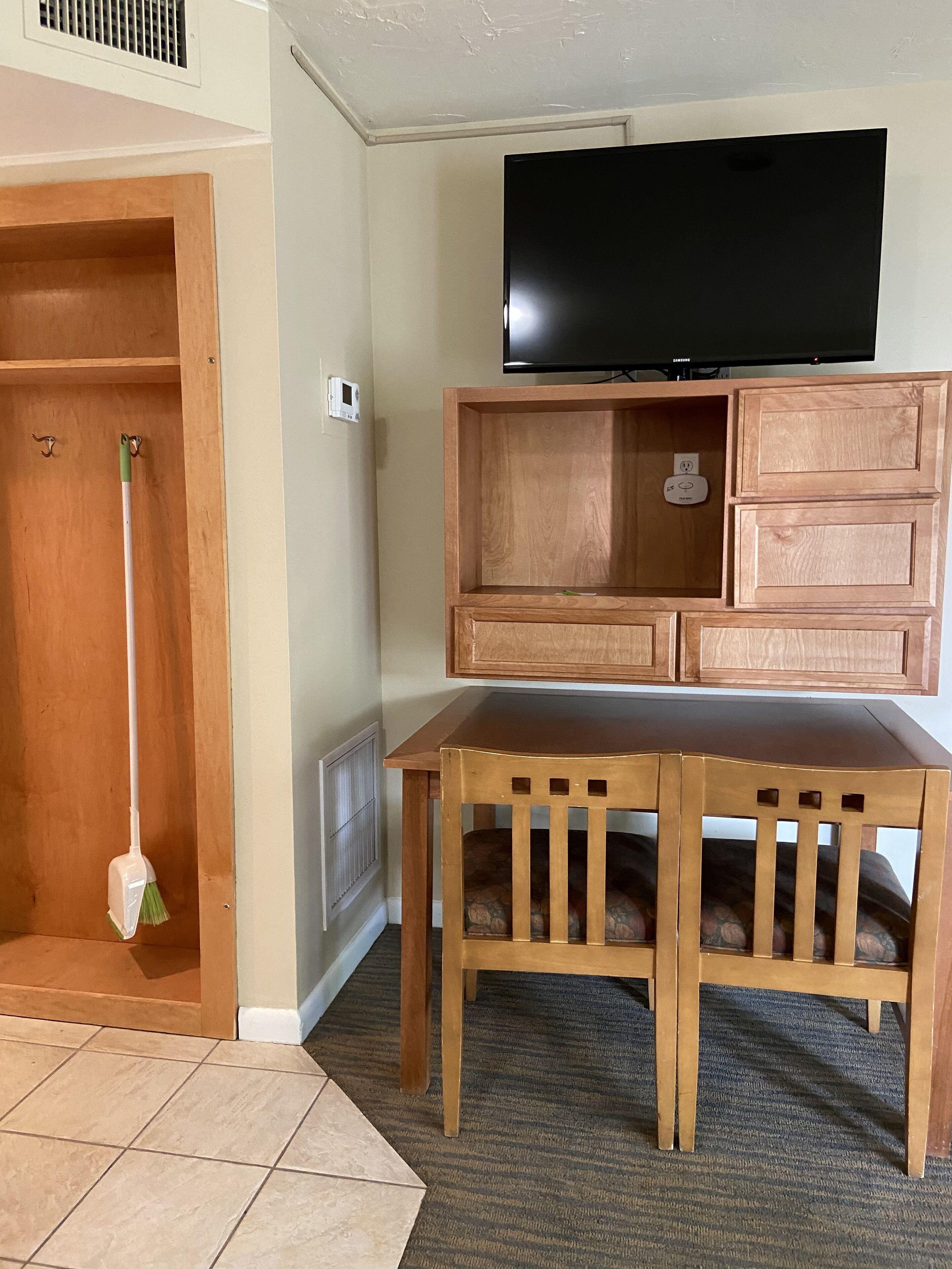 A room with a wooden table, a television mounted above it, and a door with a broom leaning against it.