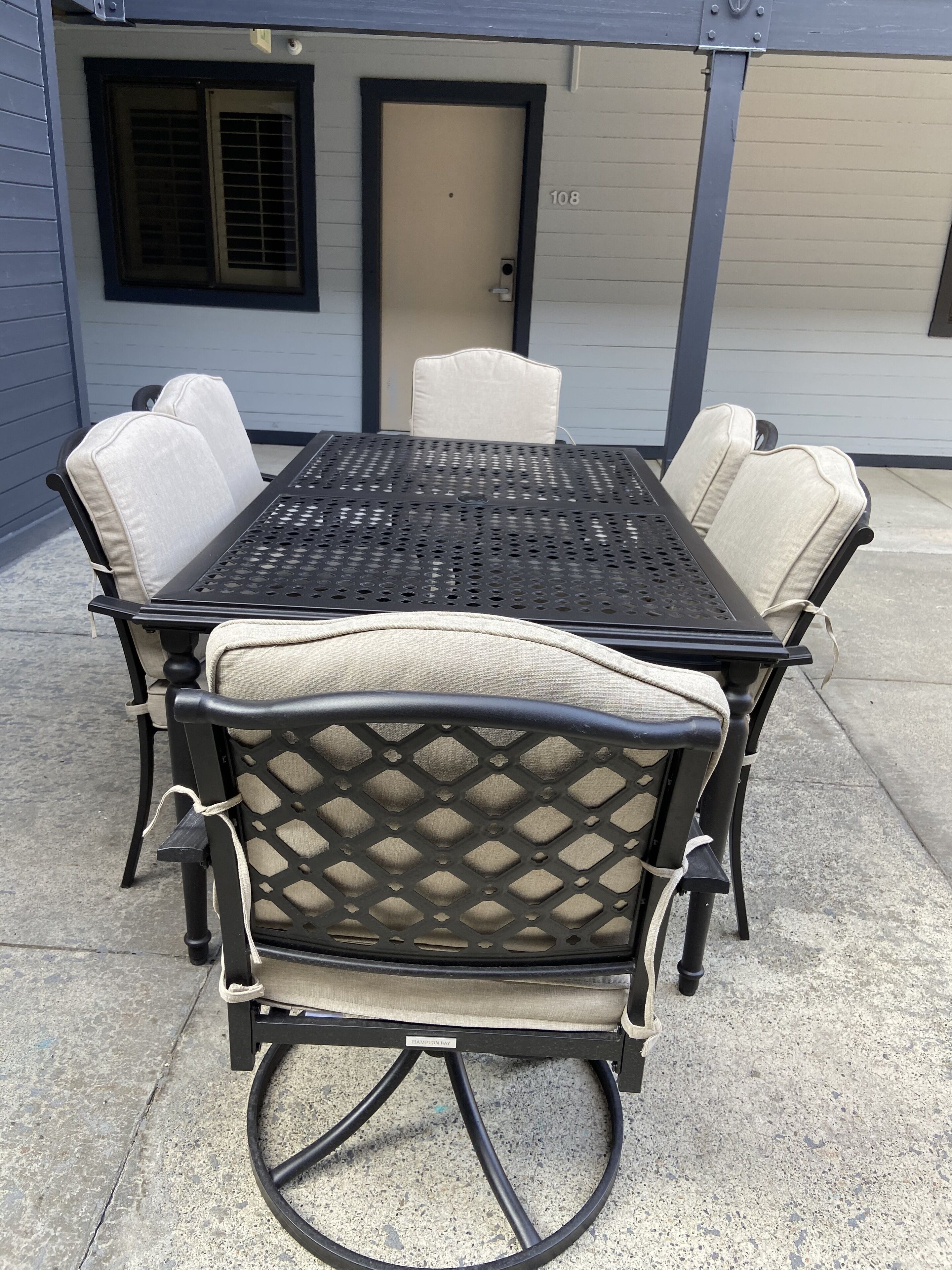 Un patio exterior con una mesa de metal y cuatro sillas, una puerta con un número y una ventana con persianas.