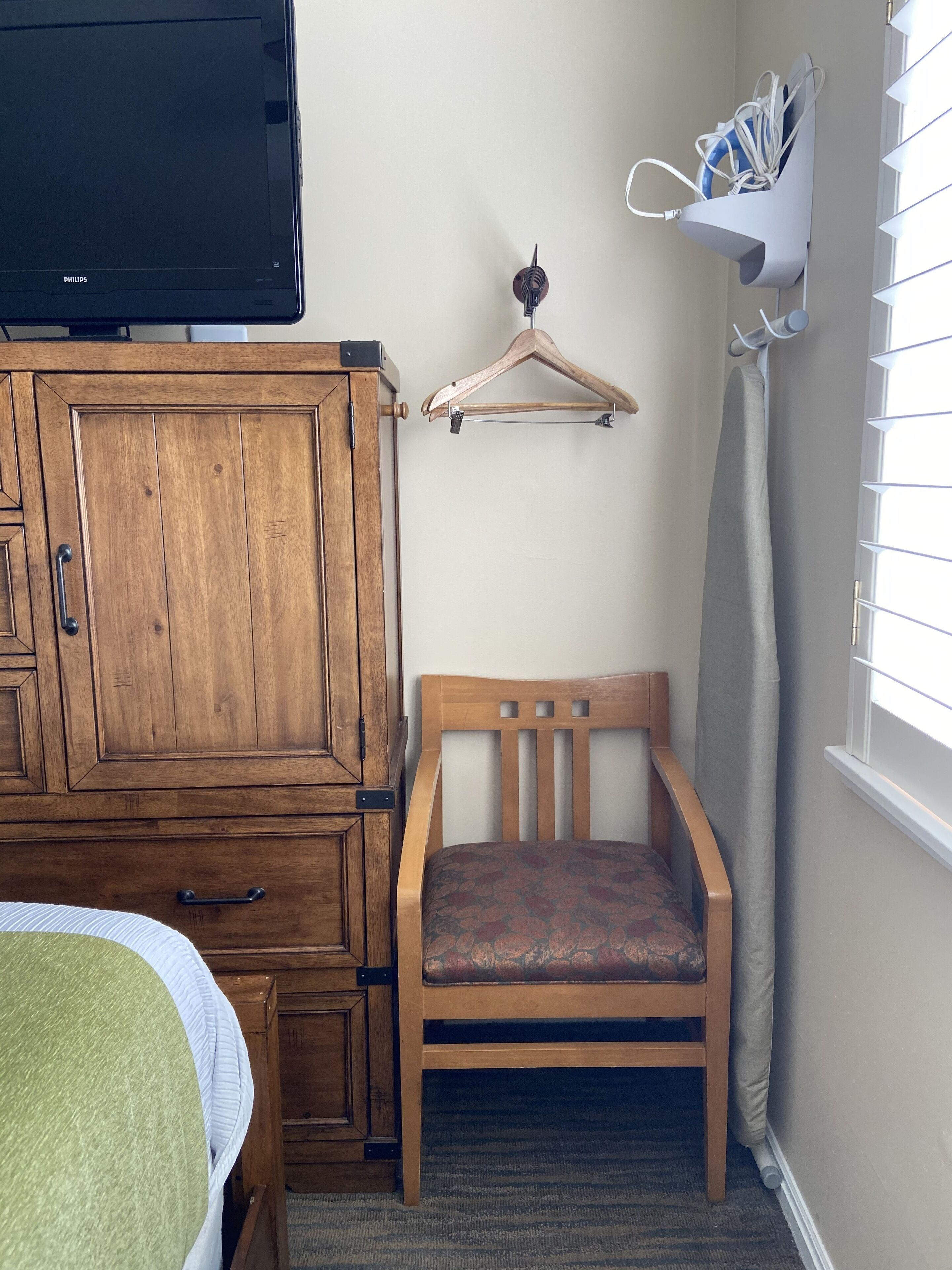 A room with a wooden cabinet, a bed with a green and white bedspread, a wooden chair, a wooden hanger, and a window with blinds.