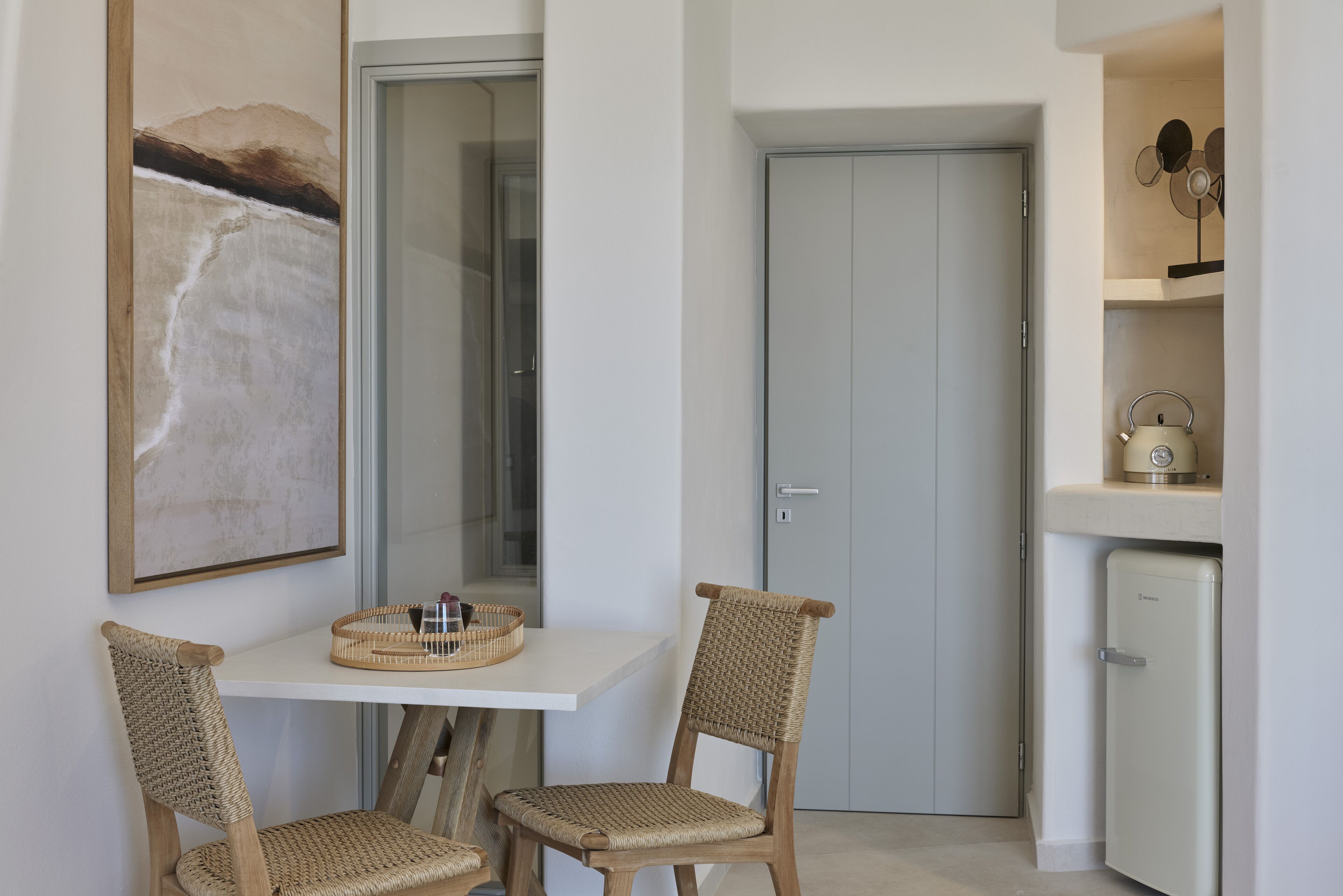 Una cocina pequeña y moderna con una mesa blanca, dos sillas de mimbre, un refrigerador y una puerta con pomo.