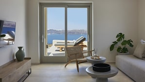 A modern living room with a large sliding glass door leading to a balcony with a view of the sea. - Apanemo Hotel (Santorini)