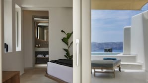 A modern bedroom with a bed, a bedside plant, and a view of a cityscape and ocean. - Apanemo Hotel (Santorini)