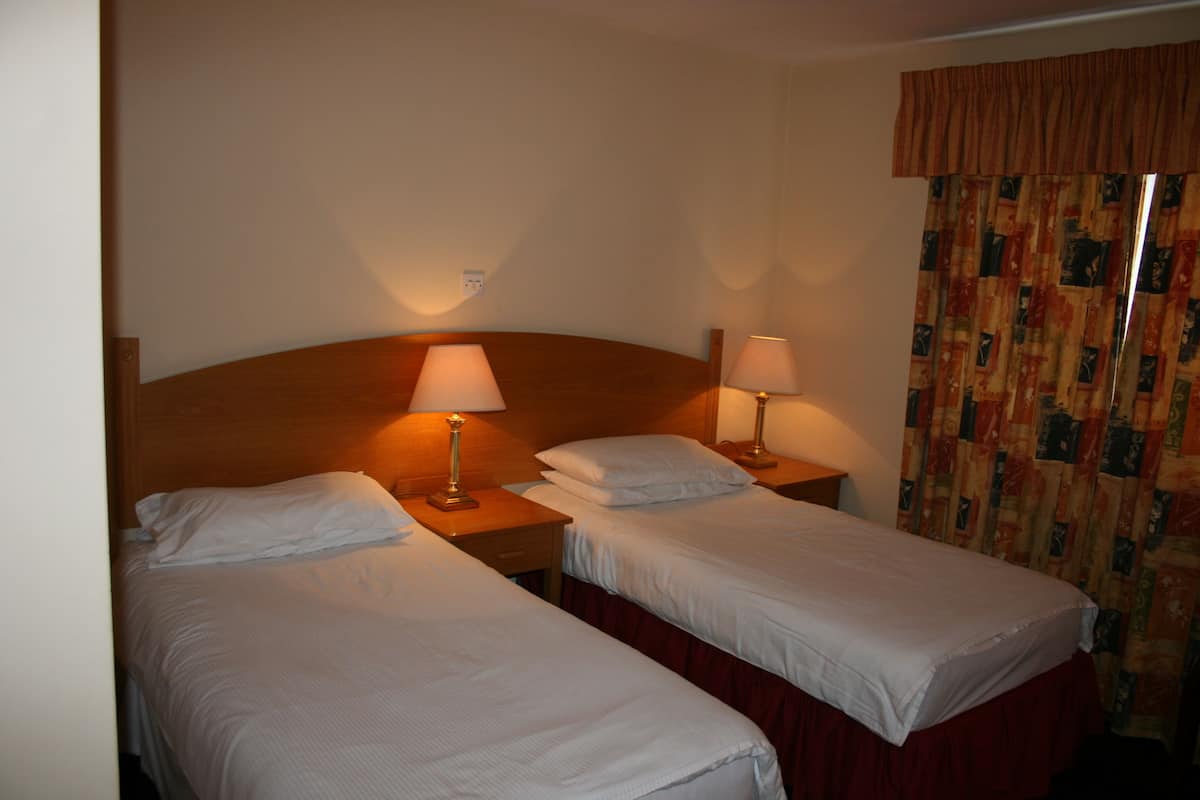 A hotel room with two single beds, each with a nightstand and a lamp. The room has a wooden headboard, a window with curtains, and a wall socket.