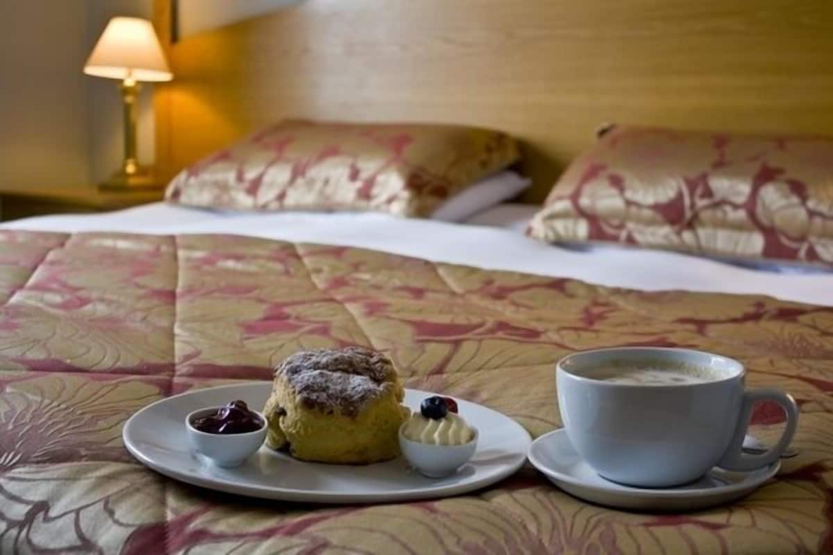 A neatly made bed with floral bedding, a cup of coffee, and a scone with jam and a blueberry on a plate.