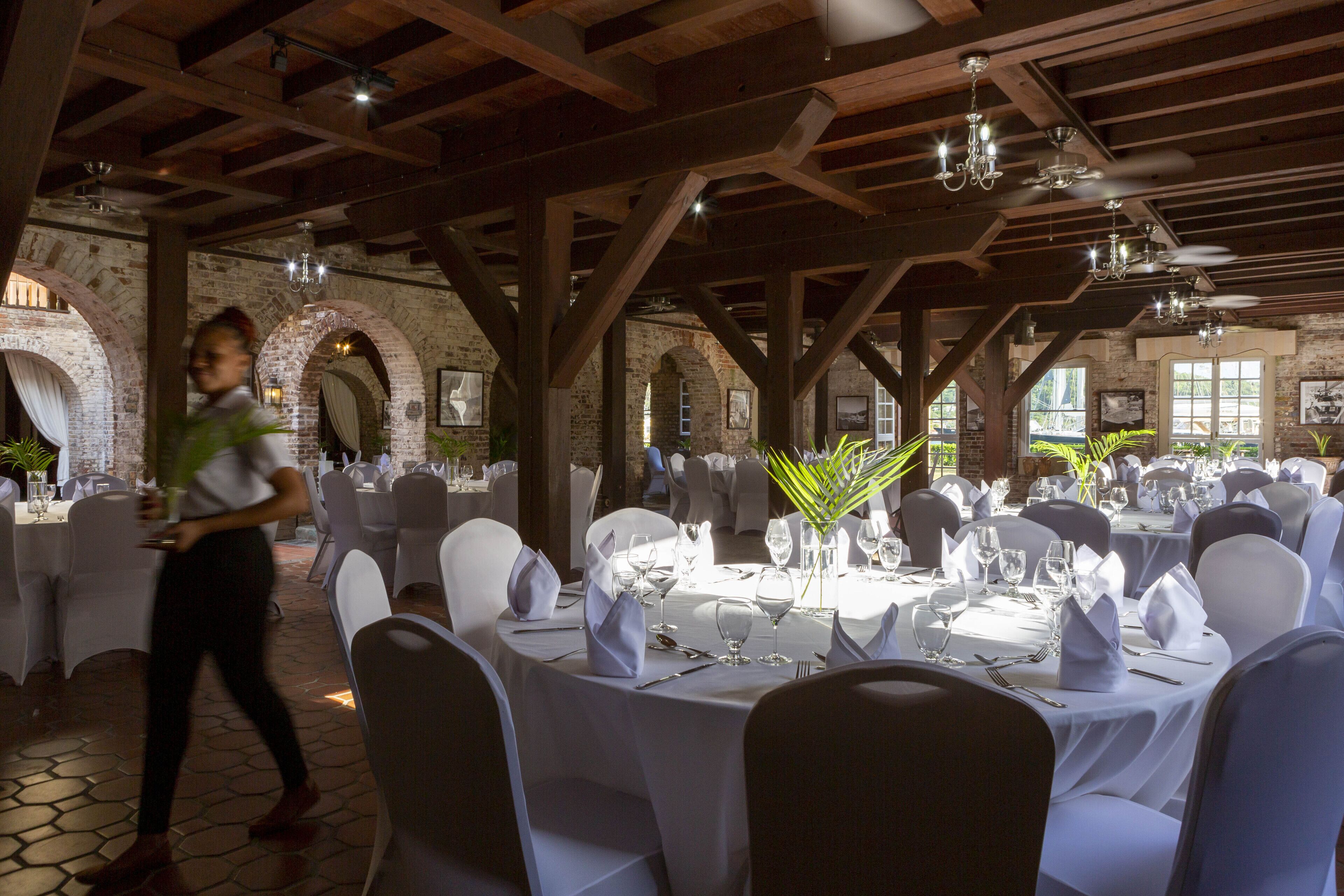 An indoor banquet hall with round tables set for dining, wooden beams on the ceiling, and brick walls.