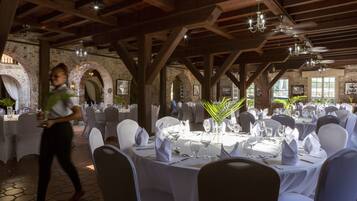 Un salón de banquetos interior con mesas redondas dispuestas para comer, vigas de madera en el techo y paredes de ladrillo.