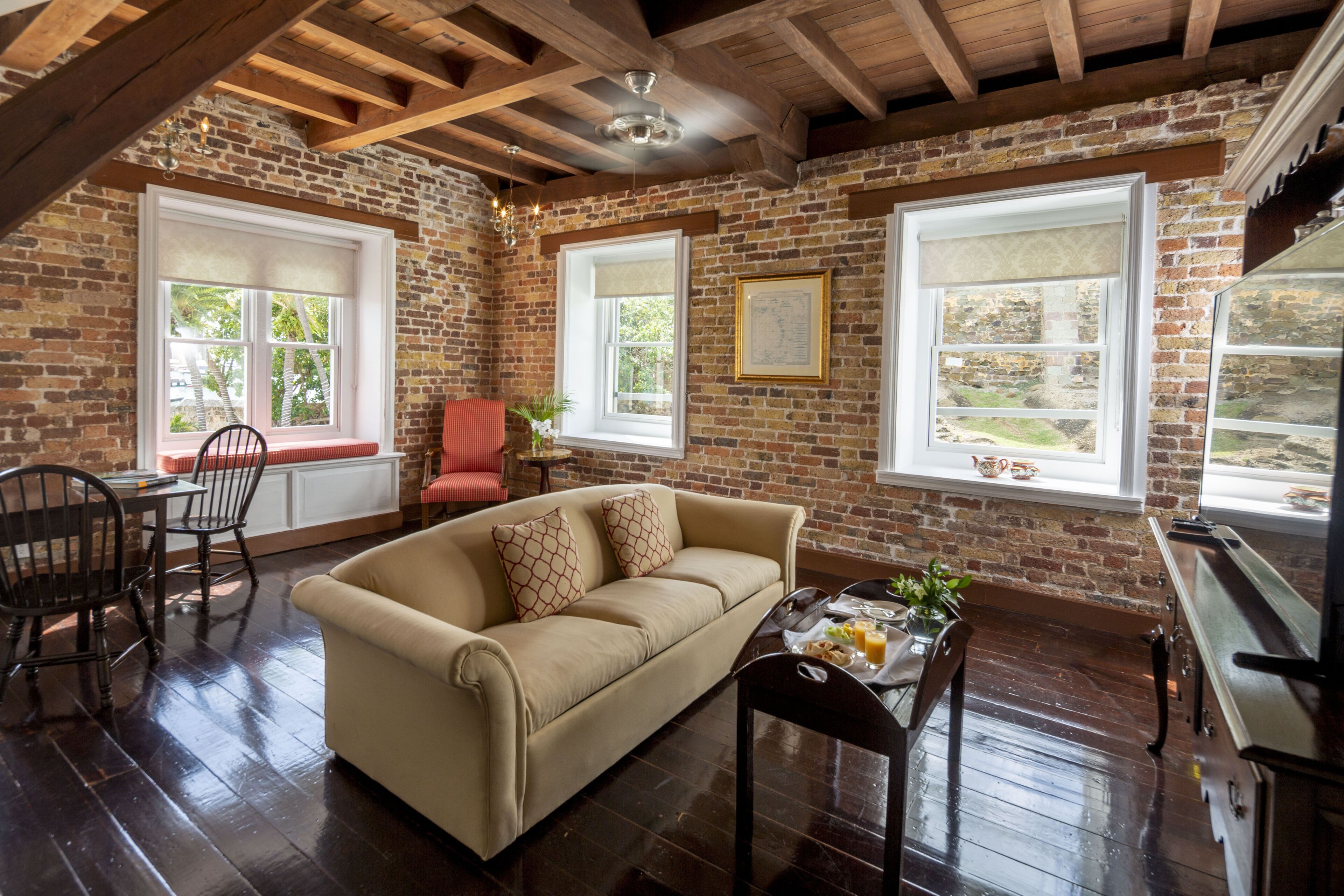 A cozy room with a brick wall, wooden beams, a beige sofa, a dining table with chairs, and a kitchen area.