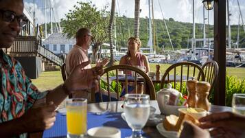 Un coin repas avec une table dressée pour quatre personnes, entourée de chaises, et en arrière-plan, une vue sur des bateaux et la verdure.
