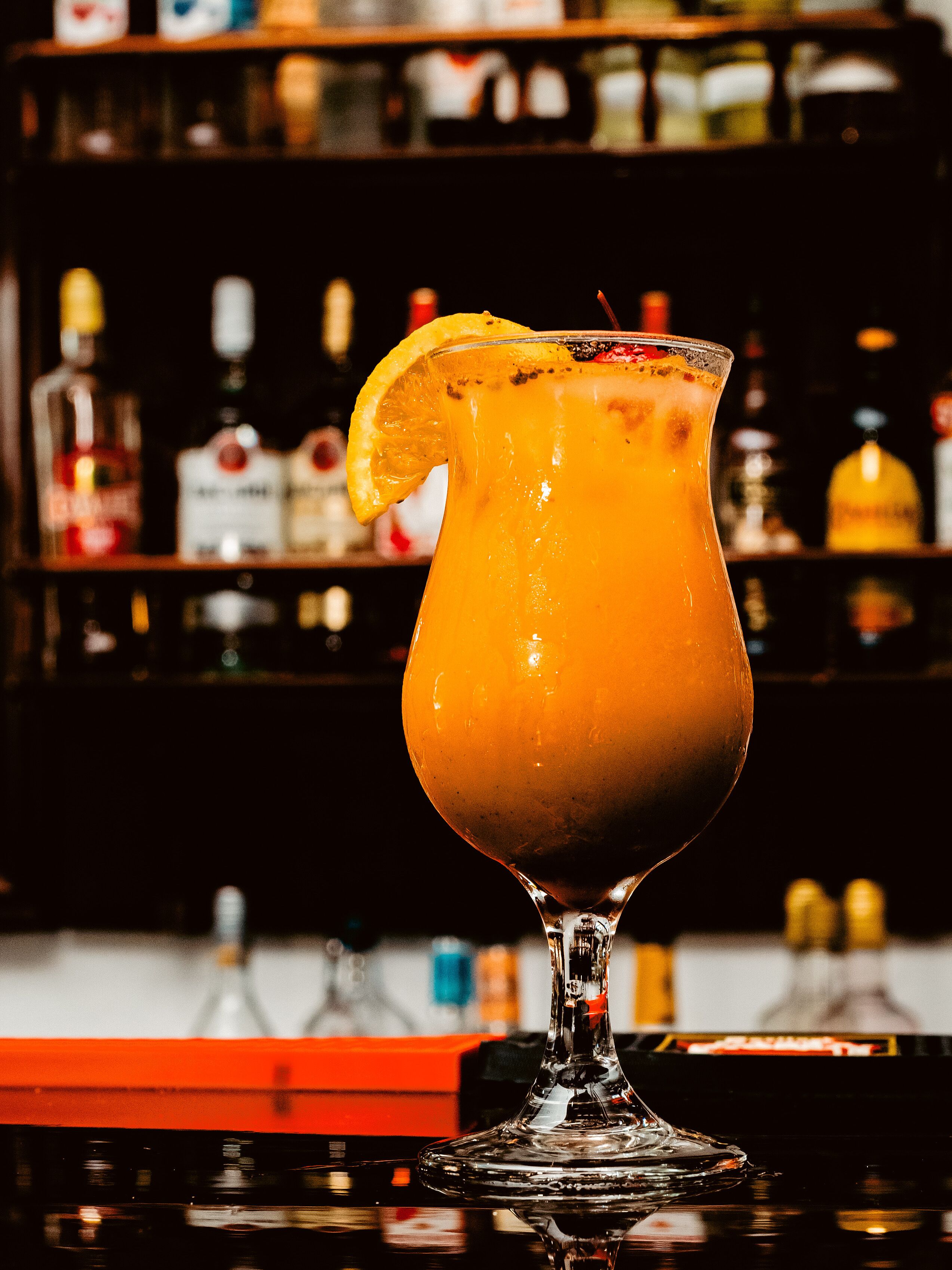 A cocktail in a glass with a lemon slice garnish, placed on a bar counter with a blurred background of shelves with bottles.
