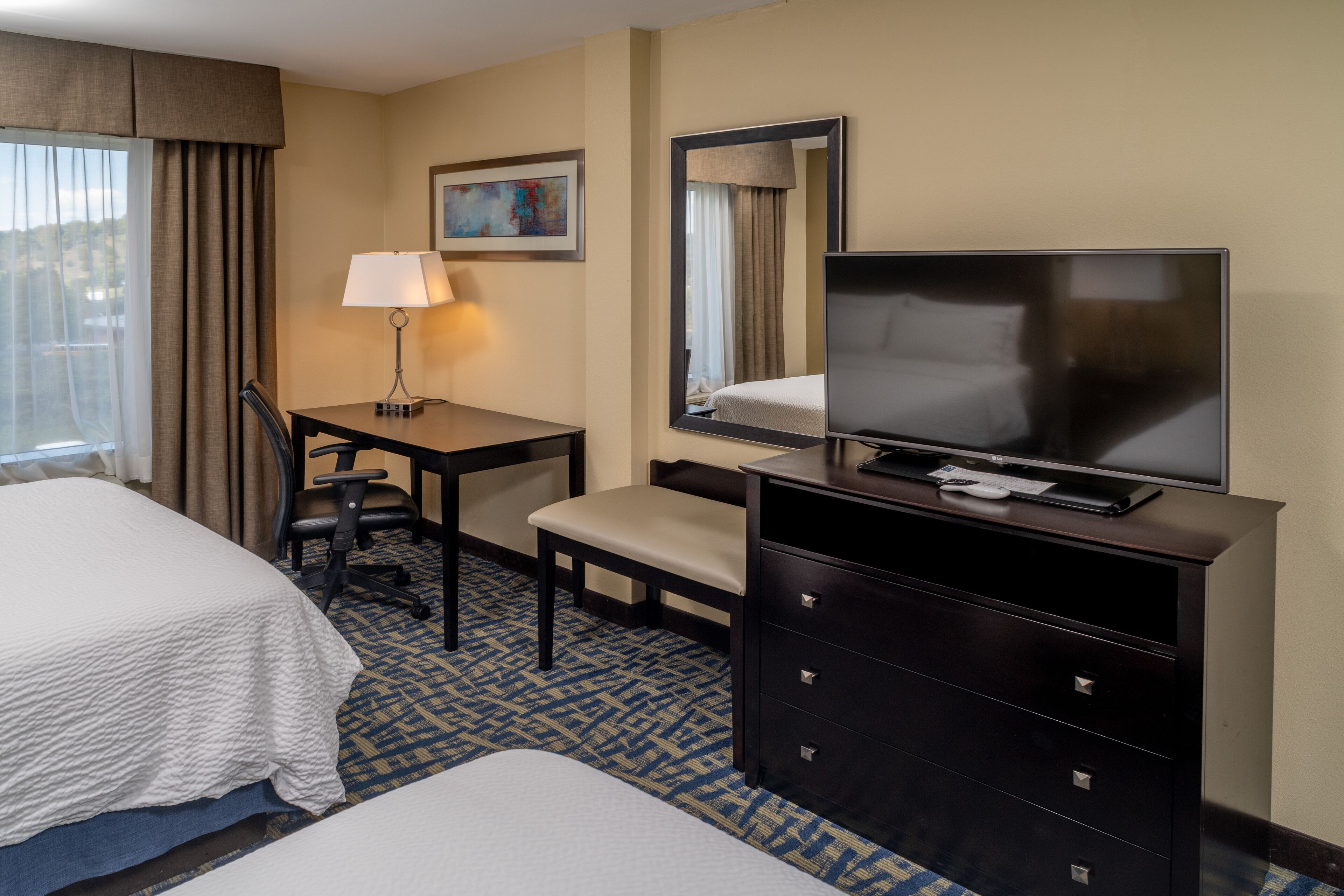 A hotel room with a bed, a desk with a chair, a television on a dresser, and a window with curtains.