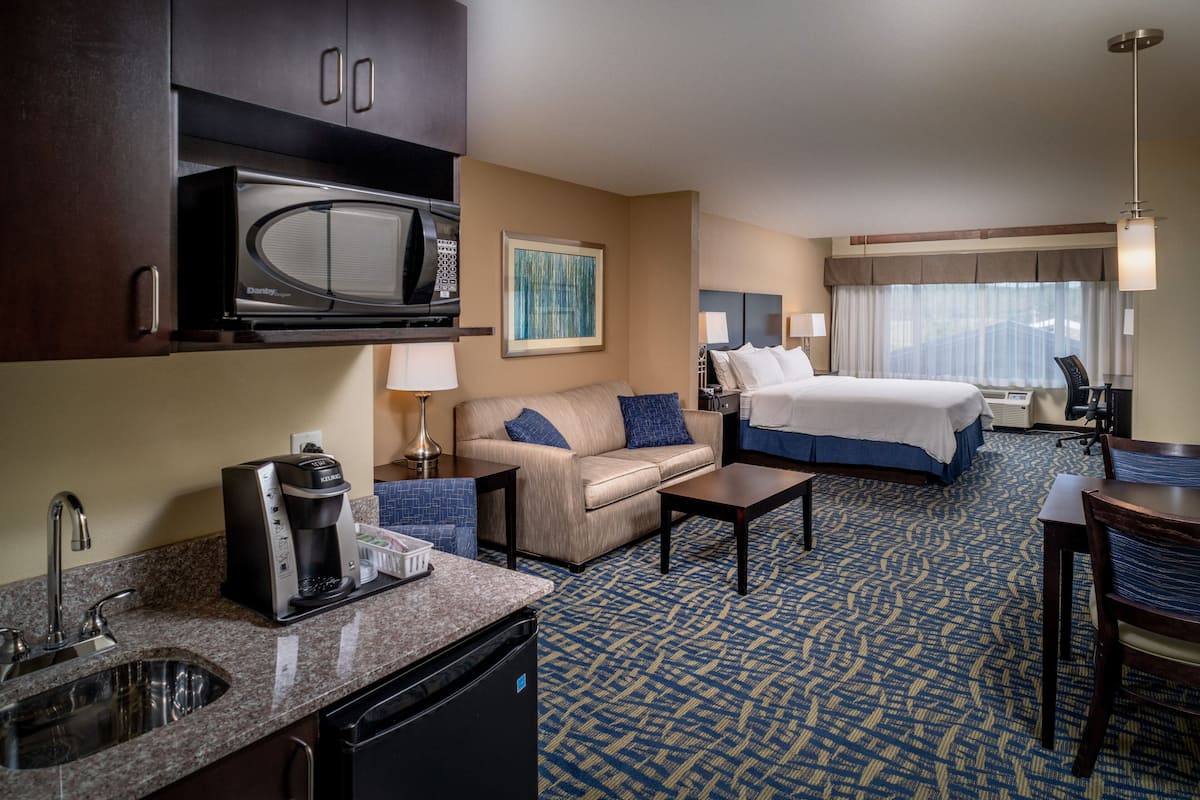 A hotel room with a kitchenette, a bed, a sofa, a desk, and a large window.