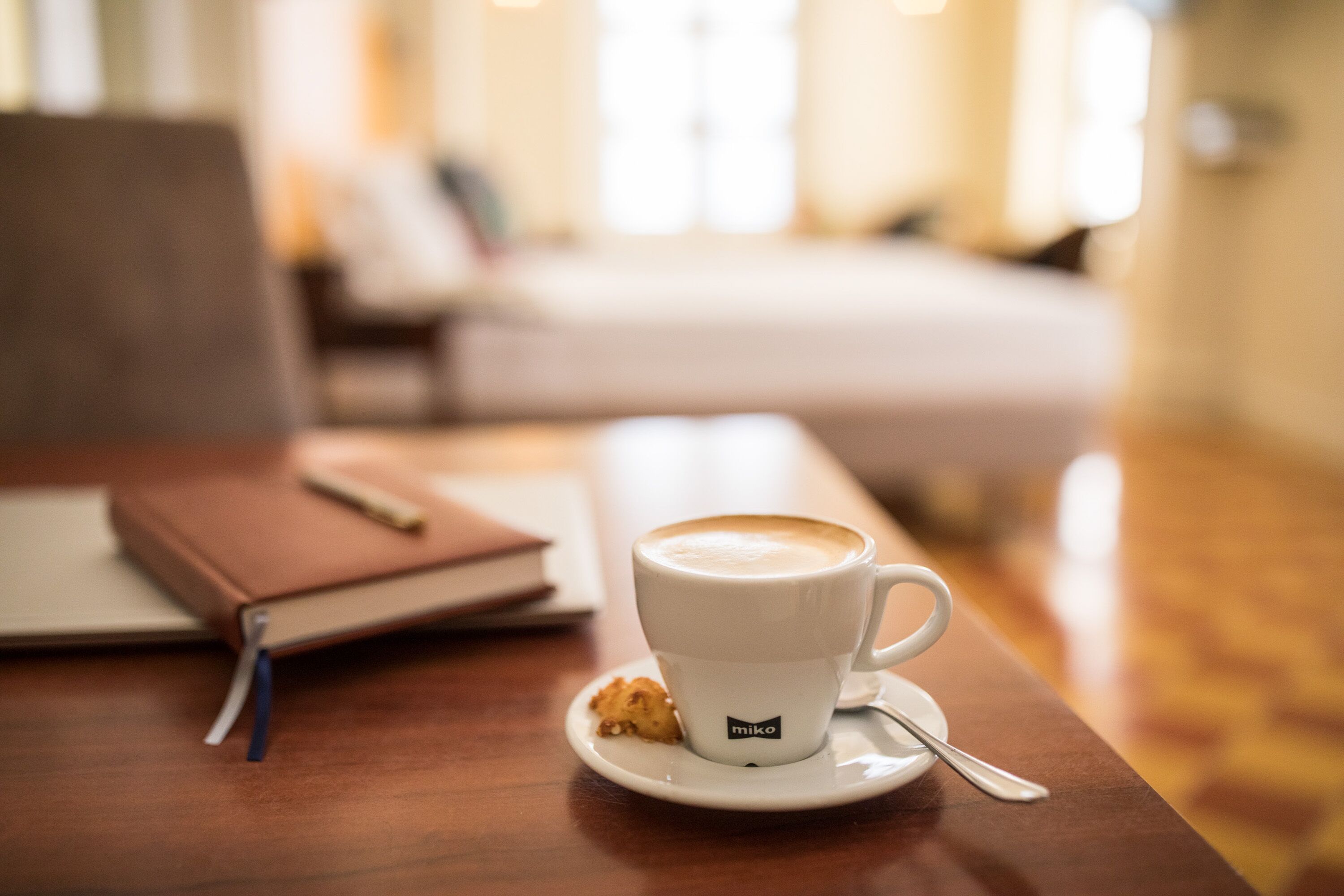 A cup of coffee on a saucer with a spoon, a notebook, and a pen on a wooden table in a room with a bed in the background.
