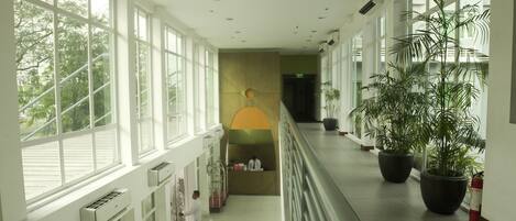 A hotel corridor with large windows, potted plants, and air conditioning units.