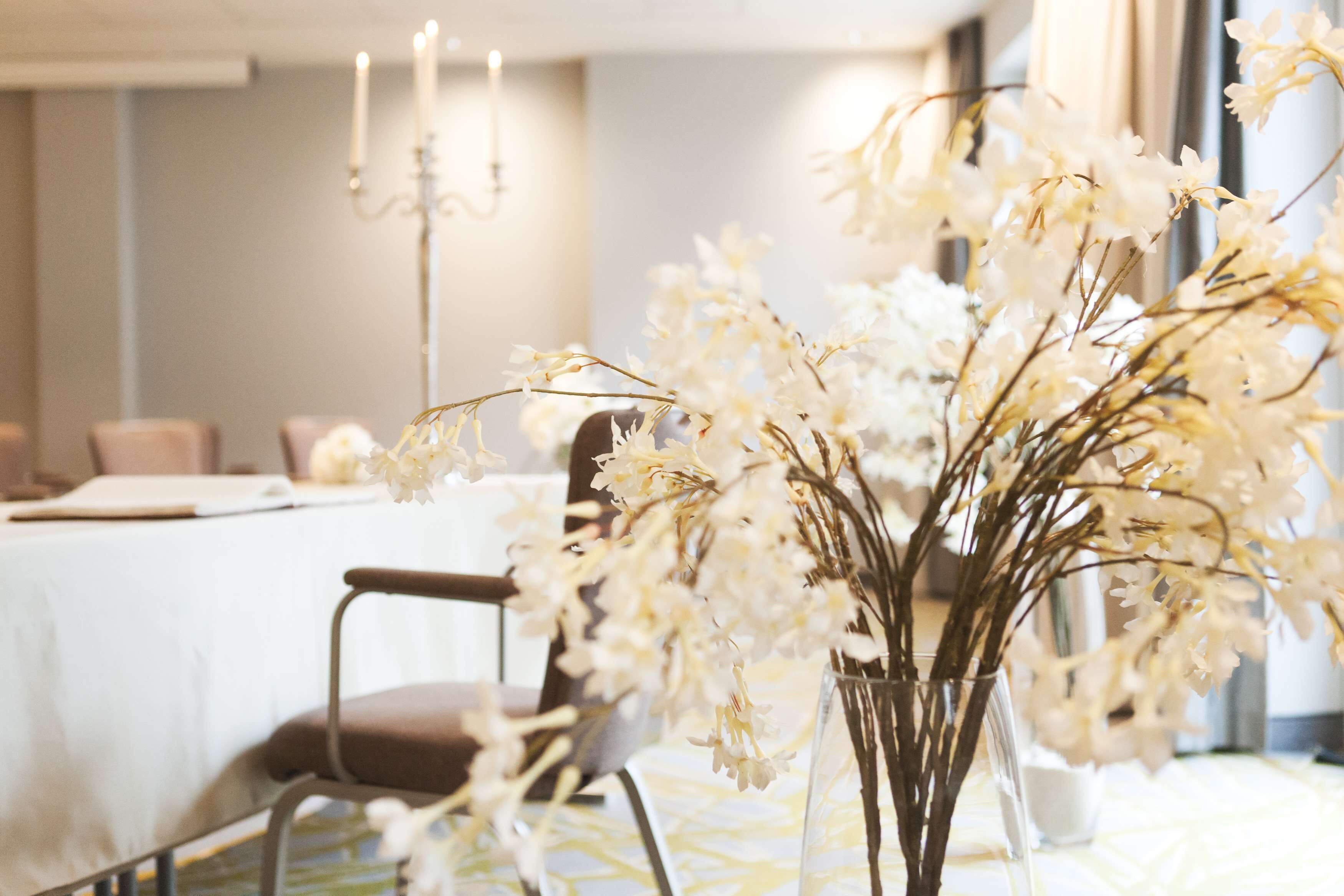 A table with a vase of white flowers, a candle, and a chair in a room with a neutral color scheme.