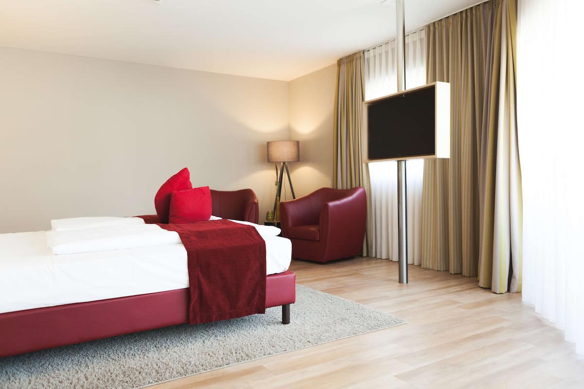 Habitación de hotel con cama, un sillón rojo, una lámpara de pie y un televisor montado en la pared.