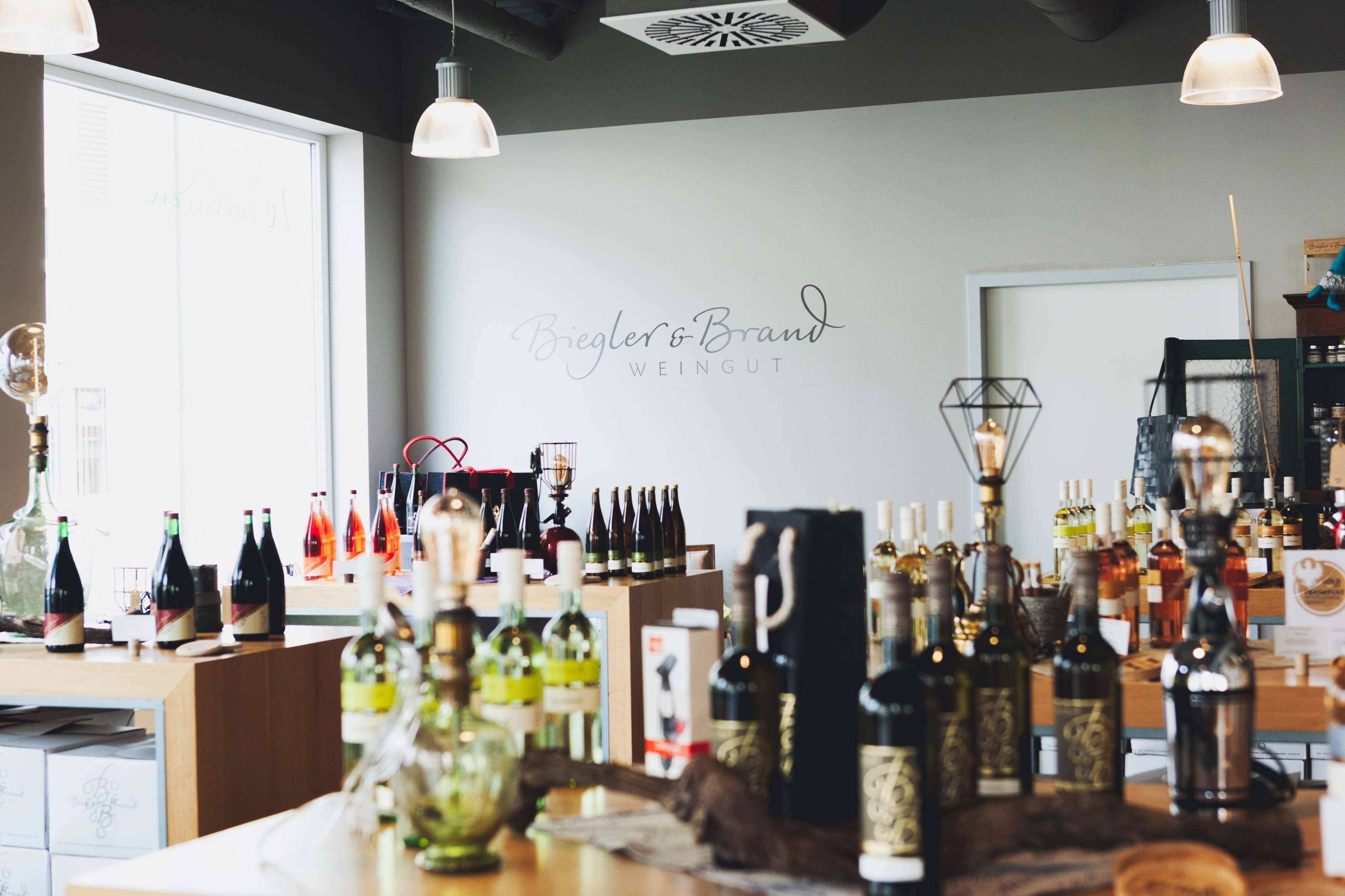 Interior de uma loja de vinhos, com diversas garrafas de vinho em exposição e um letreiro com a inscrição "Biegler & Brand WEINGUT".