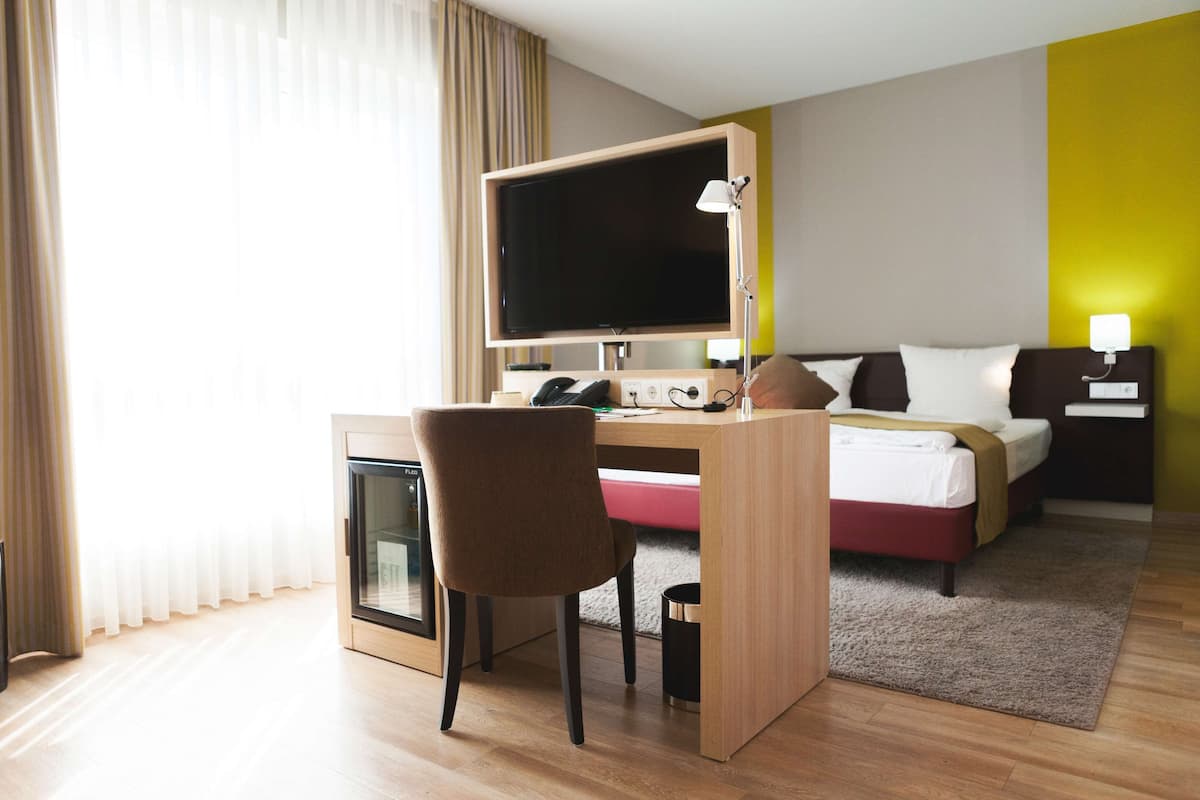 Habitación de hotel con cama, escritorio, silla, televisor, mesita de noche, lámpara y un pequeño cubo de basura.