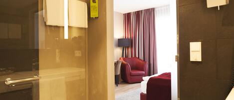 A hotel room with a glass shower door, a red chair, a small table with a lamp, and a bed with a red blanket.