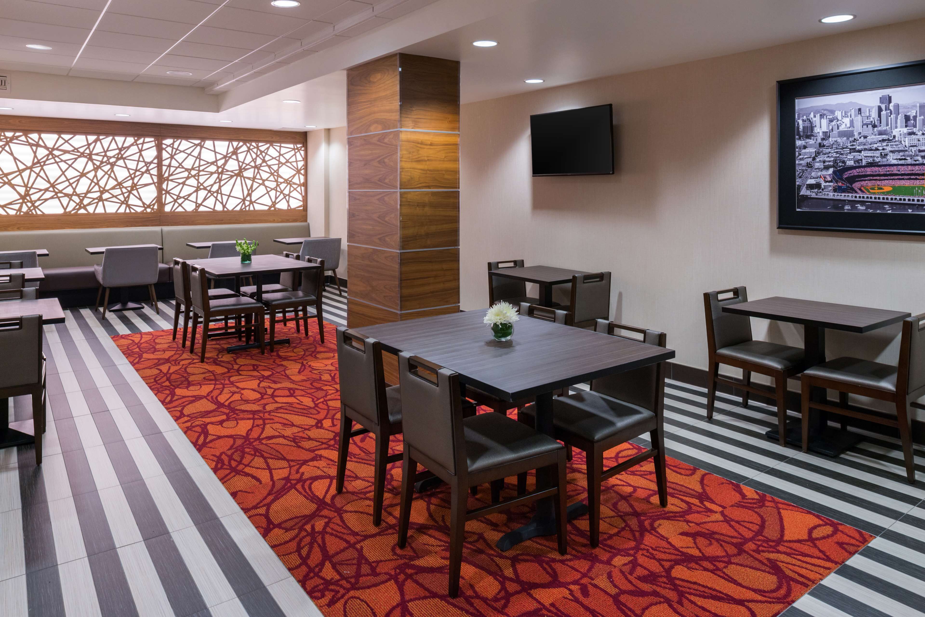 A hotel room with a large window, a flat-screen TV mounted on the wall, and a red patterned carpet.
