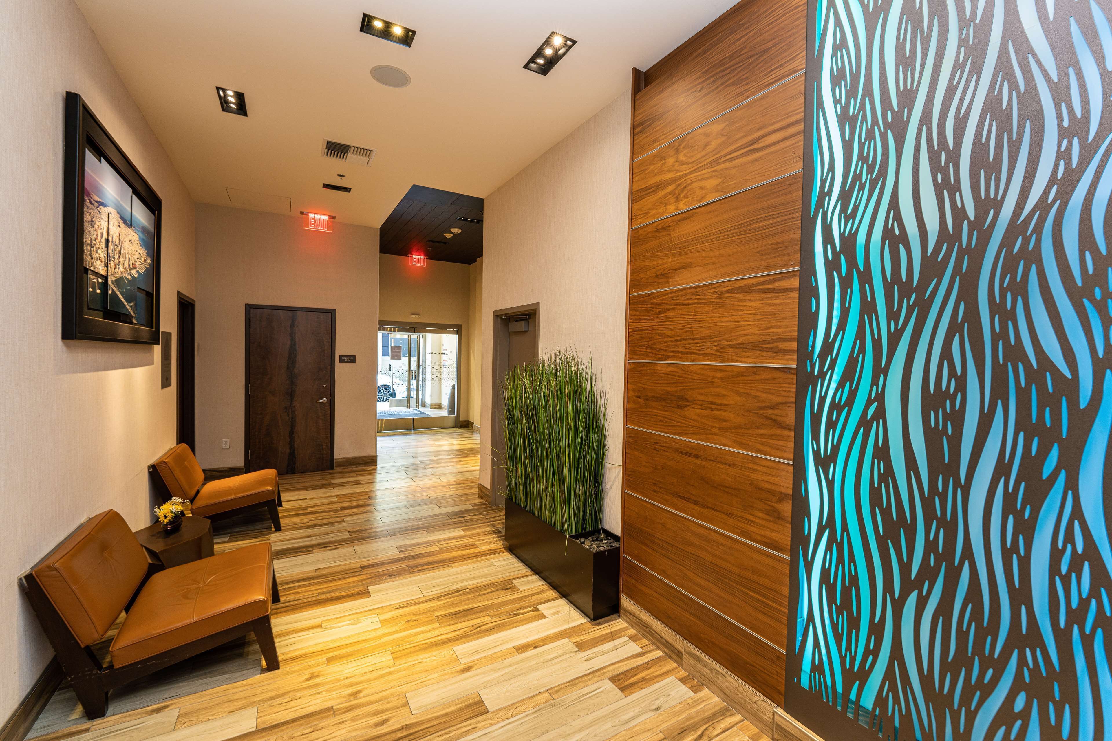 A hotel lobby with wooden flooring, a reception desk, two leather chairs, a potted plant, and a decorative wall panel.