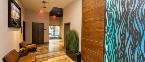 A hotel lobby with wooden flooring, a reception desk, two leather chairs, a potted plant, and a decorative wall panel.