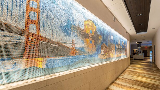A corridor with a mosaic wall depicting a bridge and cityscape, wooden flooring, and a reception area in the distance. - Hampton Inn San Francisco Downtown/Convention Center (San Francisco)