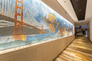 A corridor with a mosaic wall depicting a bridge and cityscape, wooden flooring, and a reception area in the distance. - Hampton Inn San Francisco Downtown/Convention Center (San Francisco)