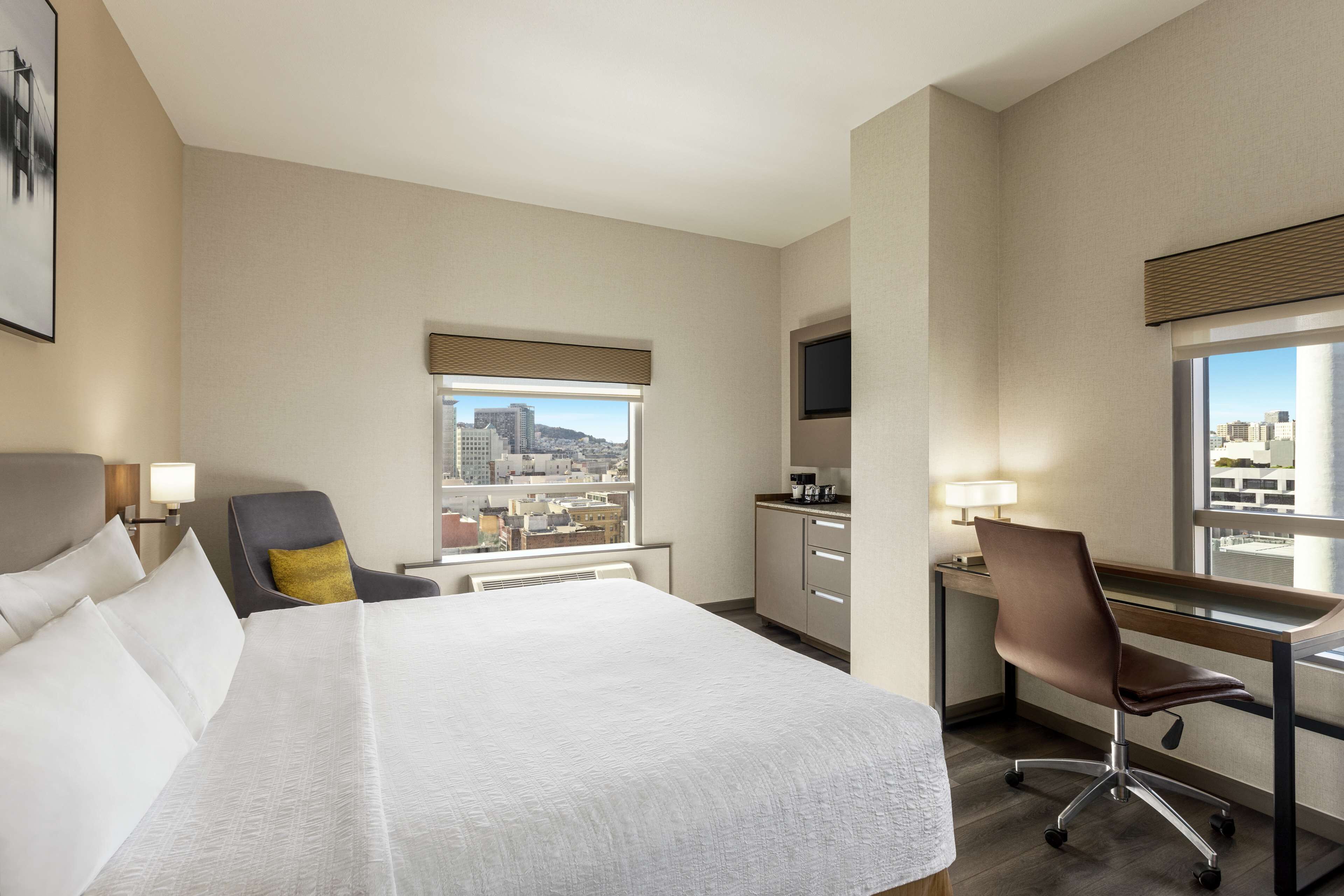 A hotel room with a large bed, a desk, a chair, a window with a city view, and a television.