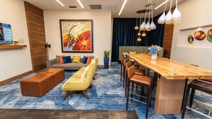 A modern hotel lobby with a wooden reception desk, a large painting, a sofa with colorful cushions, a yellow armchair, and a dining area with a wooden table and chairs. - Hampton Inn San Francisco Downtown/Convention Center (San Francisco)