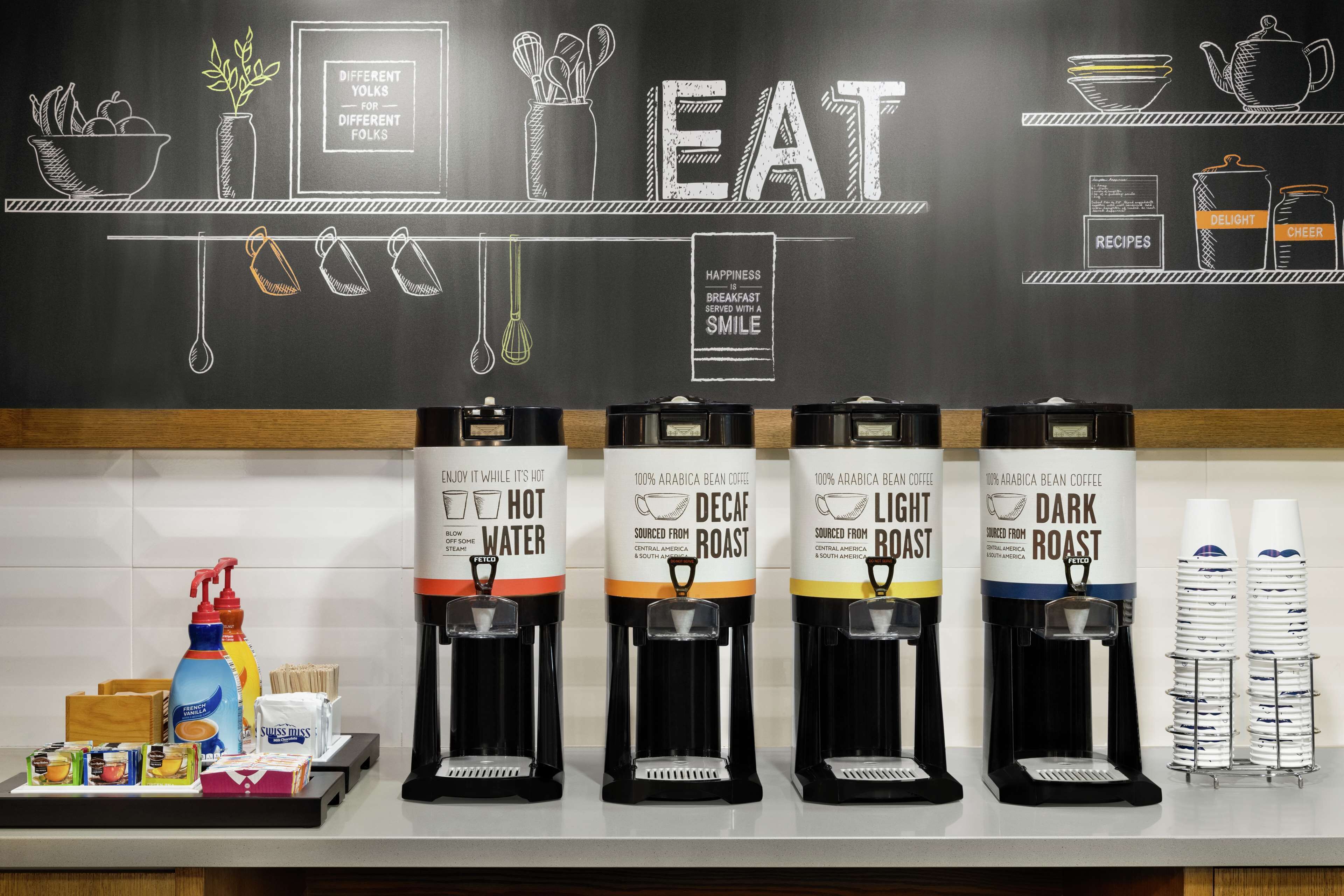 Una estación de café con cuatro dispensadores etiquetados como 'Decaf Roast', 'Light Roast', 'Dark Roast' y 'Agua Caliente'. También hay condimentos y una botella de detergente para platos en la encimera.