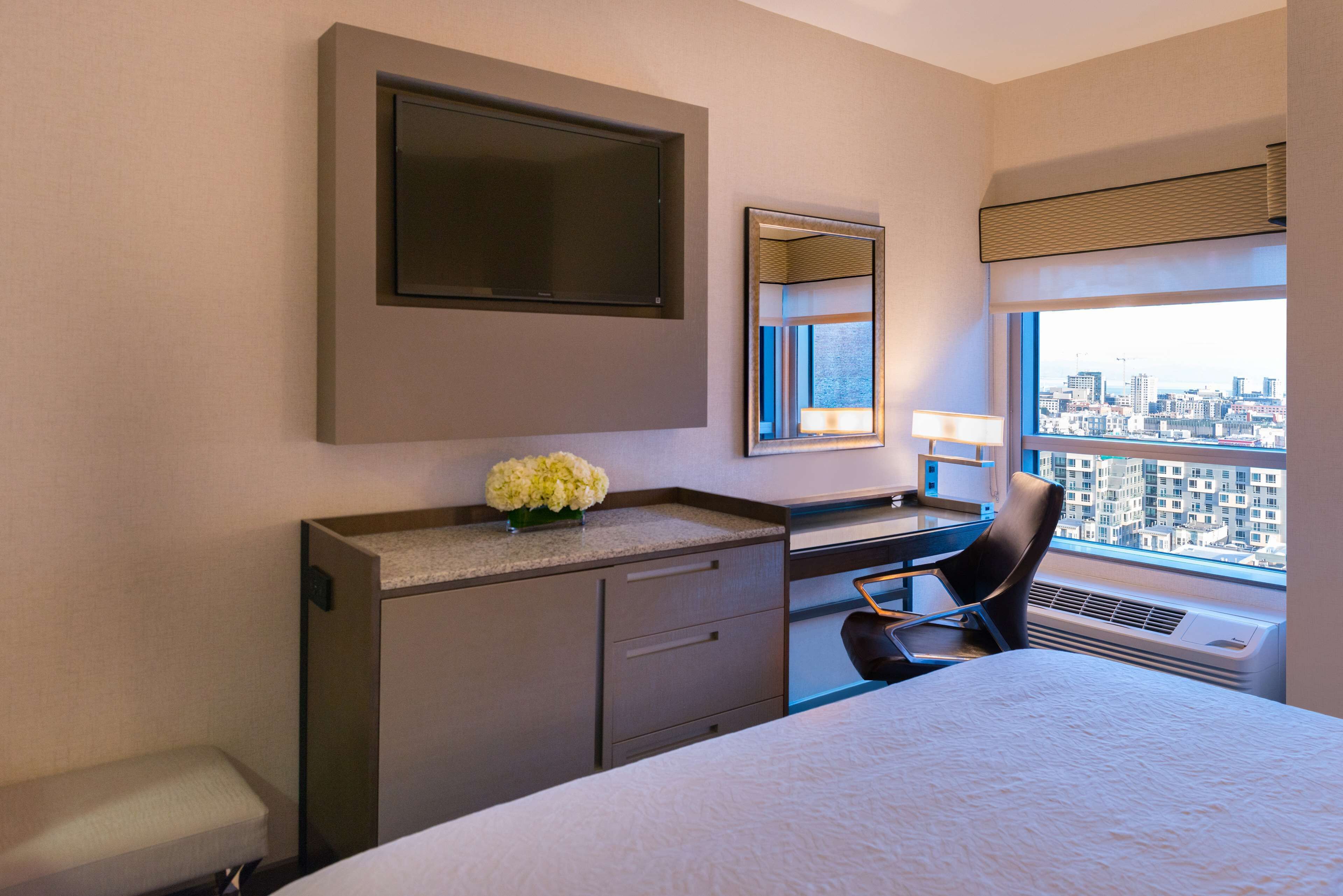 A hotel room with a bed, a desk with a chair, a dresser, a mirror, and a window with a city view.